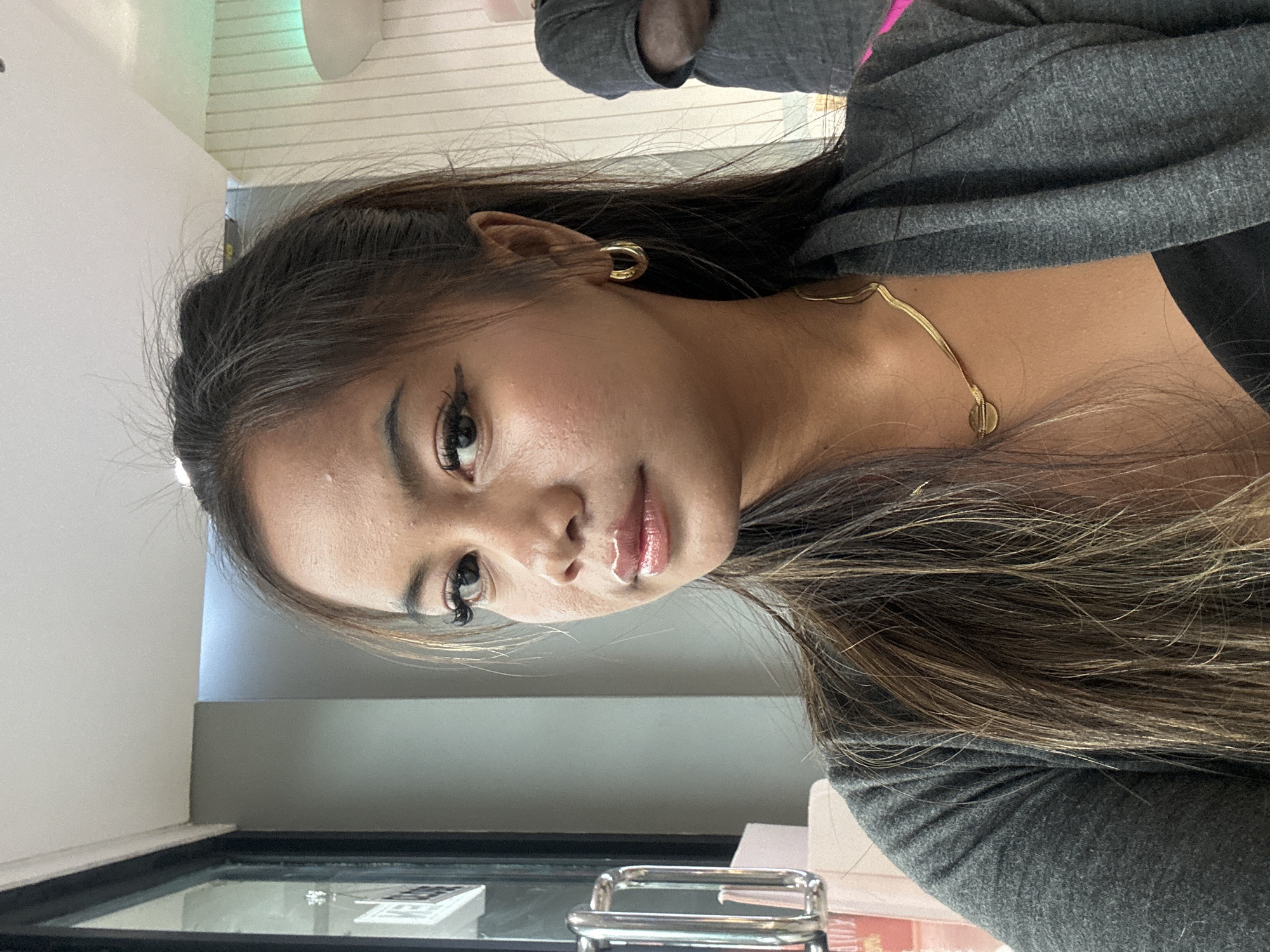 A person with long, brown hair and light skin is wearing a gray sweatshirt. They are also wearing gold hoop earrings and a delicate gold necklace. The person is looking towards the camera with a neutral expression. The background includes a gray wall and a window with a black frame. The location is inside, possibly a café or restaurant in North Jakarta, Indonesia. A metal object is visible near the bottom of the frame on top of the table.