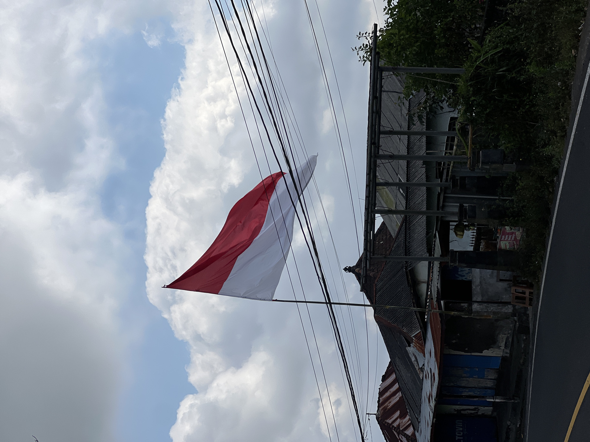 An Indonesian flag, red and white, flies from a tall pole against a cloudy blue sky in Kintamani, Indonesia. Below the flag, a row of aging buildings with corrugated metal roofs and weathered facades lines a road. Power lines crisscross the sky above the structures. The buildings appear to be small shops or residences, some with peeling paint and visible rust. Lush green foliage encroaches on the right side of the frame, bordering the buildings. The road in the foreground is dark asphalt with a yellow dividing line. The scene conveys a sense of everyday life in a tropical location.