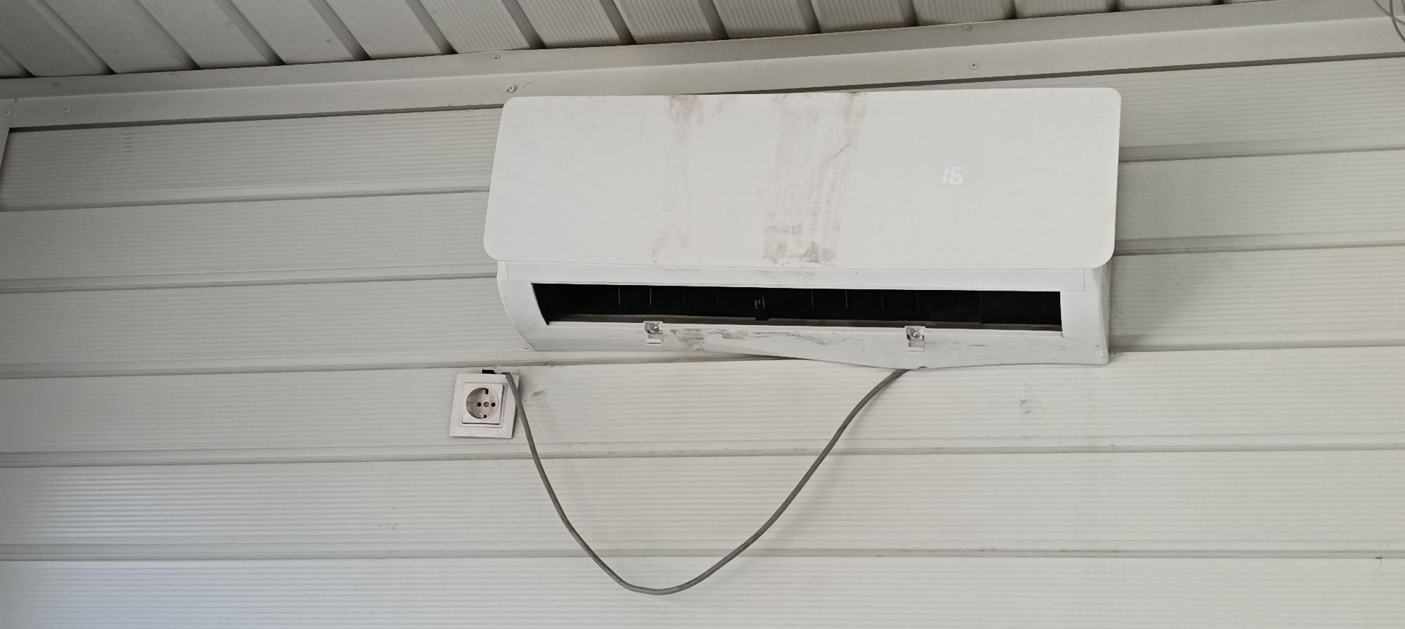The image shows a close-up of a white, split air conditioning unit mounted on a white, horizontally ribbed wall. The unit has a digital display showing the number "16". A gray power cable extends from the unit and plugs into a white electrical socket mounted on the wall below and to the left of the air conditioner. The wall appears to be made of siding, and the ceiling above is also visible, composed of white, ribbed material. The air conditioning unit and the surrounding wall show signs of dust and staining. The scene is well-lit, suggesting it might be daytime. The location is identified as Qarağandı city administration, Kazakhstan. There are no people or animals visible in the image.