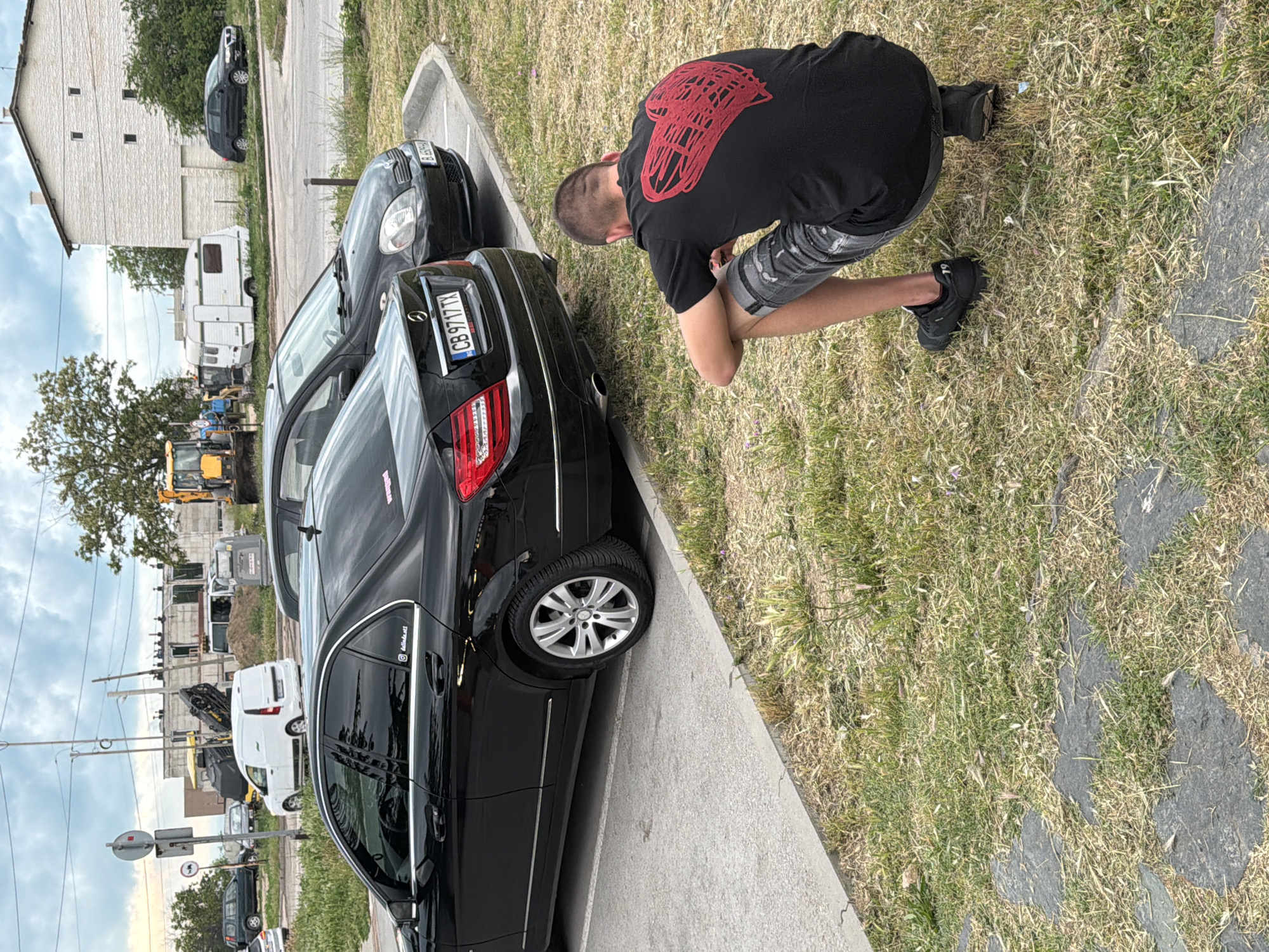 The image depicts an outdoor scene in Varna, Bulgaria, centered around a young man kneeling beside a black Mercedes-Benz sedan. The man is wearing a black T-shirt with a large red, abstract design on the back, gray patterned shorts, and black sneakers. His posture suggests he is examining something low to the ground or possibly taking a photo.
The black Mercedes-Benz is parked on a concrete surface next to a grassy area. Its rear license plate, "CB 9717 TX", is clearly visible, as is an Instagram handle "@dmds.atl" on its rear side window. Another dark car is parked directly in front of the Mercedes, with a partially visible license plate starting with "B 639".
The background reveals a mix of urban and semi-urban elements. A white multi-story building stands beyond the parked cars. Further in the distance, several vehicles are visible, including a white caravan, a white van, and construction equipment such as an excavator. The sky is overcast, indicating a cloudy daytime setting. The ground near the man is covered in sparse, dry grass with some darker stone or concrete elements.