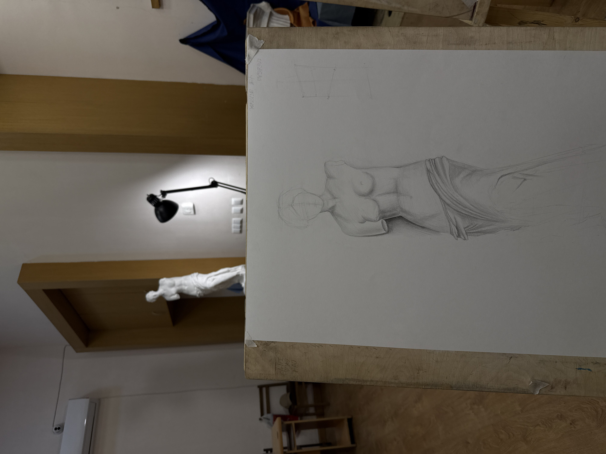 A well-lit interior space contains a drawing of a female torso draped in fabric. The drawing is displayed on a wooden drawing board. Additional light source is a lamp. A sculpture is situated in a wooden display area. The background includes wall-mounted fixtures and furniture.
Based on the text visible on the drawing board, it can be inferred that the drawing was created in Yerevan, Armenia.