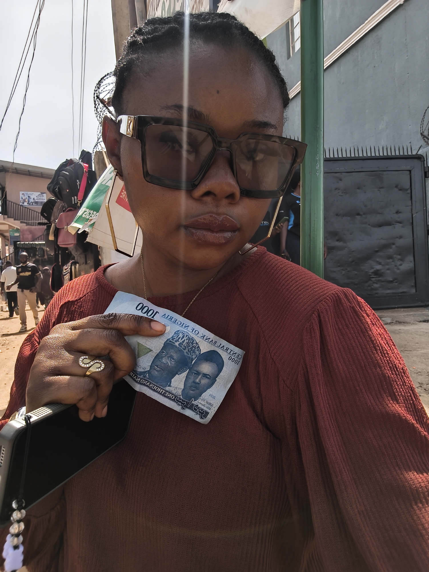 A medium close-up, eye-level shot features a person, likely female, with dark skin, posing for a photo outdoors in what appears to be a vibrant urban or market setting in Jos, Nigeria. The individual is positioned in the foreground, looking directly at the camera with a neutral expression. They wear large, dark-framed sunglasses with gold accents on the temples, a reddish-brown ribbed long-sleeved top, and a thin gold chain necklace. Their hair is styled in dark braids.
In their left hand, the person holds a black smartphone and a 1000 Nigerian Naira banknote. The banknote is presented with its reverse side facing the viewer, showing the reversed text "AIRAN DNASUOHT ENO" (ONE THOUSAND NAIRA), "AIREGIH FO KNAB LARTNEC" (CENTRAL BANK OF NIGERIA), and the numeral "0001" (1000). The portraits of two distinguished Nigerian figures are visible on the note. A prominent gold ring featuring a stylized 'S' shape adorned with white stones is worn on their left ring finger. Their lips appear glossy.
The background on the left side of the image depicts a busy street scene under a bright sky, with blurred figures of people walking and market stalls or shops displaying various bags. Overhead, electrical wires are visible. On the right, a vertical green pole stands beside a plain grey building wall and a dark, spiked metal gate. The bright sunlight suggests it is daytime, casting clear reflections on the sunglasses. The scene captures a moment from daily life in an urban Nigerian environment.