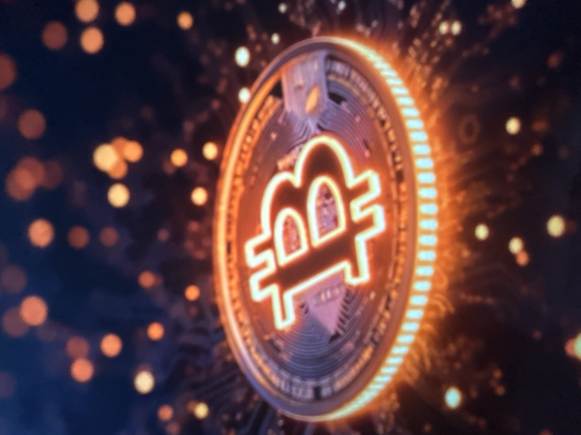 The image shows a close-up of a Bitcoin cryptocurrency coin, glowing with orange and white light, set against a dark, bokeh-filled background. The coin is centrally located and appears to be the primary subject. The background consists of out-of-focus circular lights in shades of orange and yellow, suggesting a digital or abstract environment. The Bitcoin logo, a stylized "B," is clearly visible on the coin's surface. There is no indication of any specific location, including Gelendzhik, Russia, nor are there any people, actions, or interactions depicted. The image is purely symbolic of cryptocurrency.