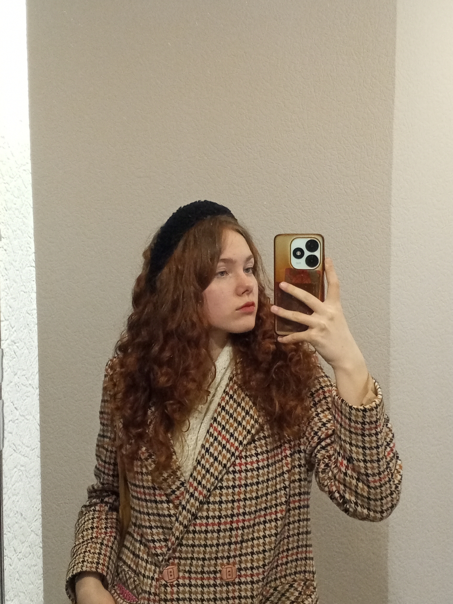 A young woman with reddish-brown curly hair and a black headband is taking a selfie with a smartphone. She is wearing a houndstooth blazer over a white knit top. The background is a plain beige wall. The photo appears to be taken indoors. The location, Chaikovsky, Russia, is specified based on external information and not evident from the image itself.
