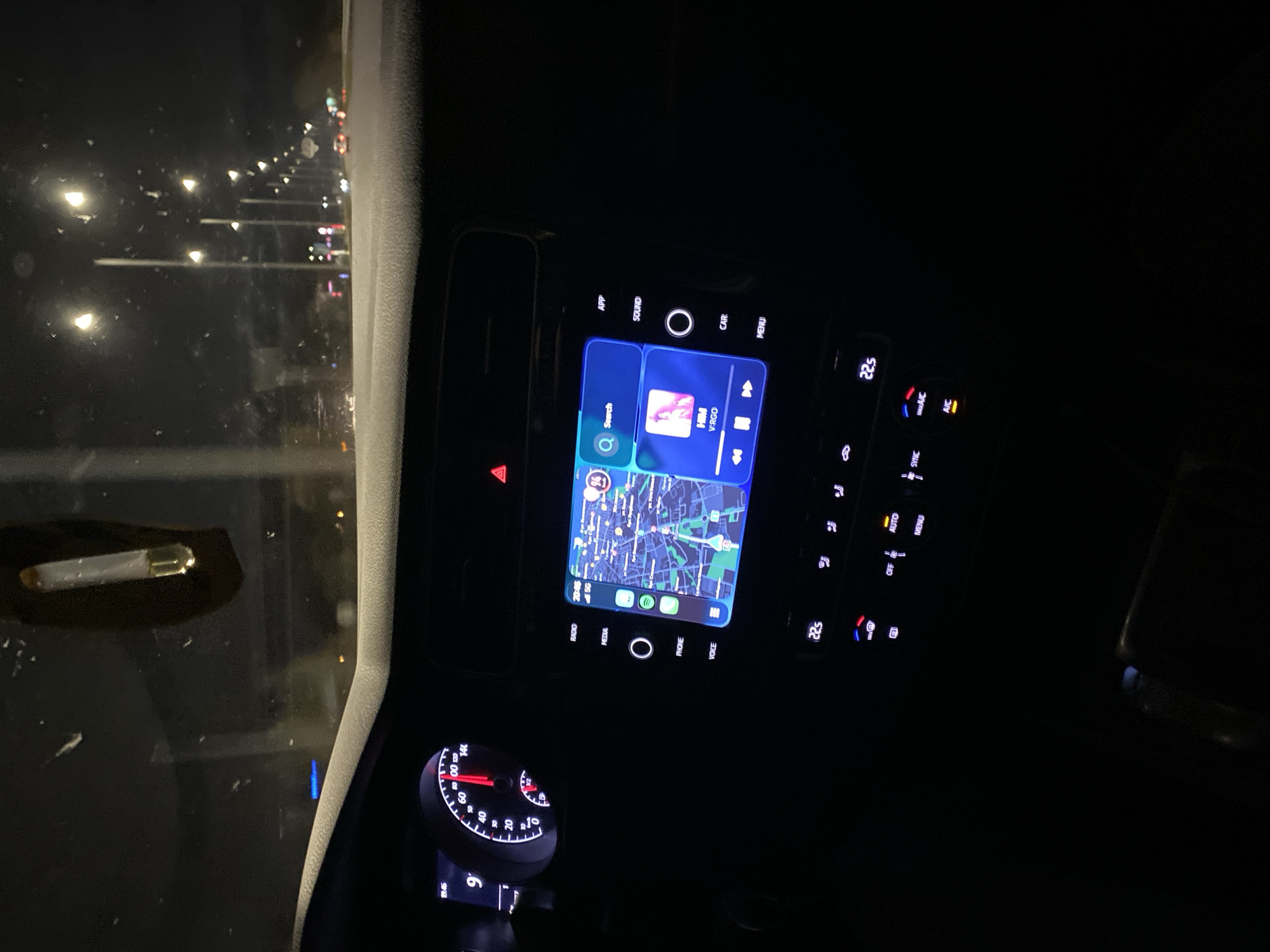 The image appears to be taken from inside a car at night. The dashboard is visible, displaying a GPS navigation system with a map. The speedometer shows a speed of around 0 km/h. The display also indicates that it is 20:46 (8:46 PM). On the screen of the infotainment system, a navigation screen is showing a map of what appears to be a city. Based on the GPS navigation screen, it seems that the driver is located in Varna, Bulgaria. The display also indicates that music is playing. Other dashboard elements include climate control buttons, a hazard light button, and other controls. Outside the window, street lights and other vehicles are visible. There is an object near the windshield that appears to be a cigarrette.