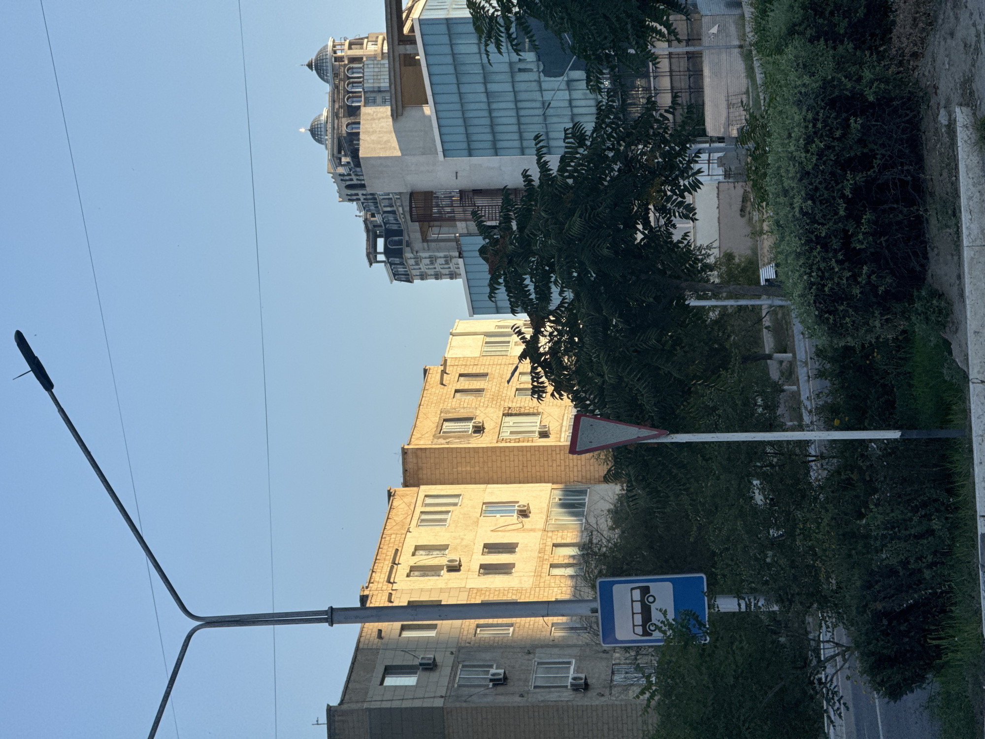 The image shows a street scene in Aktau, Kazakhstan, during daylight. In the foreground, a sidewalk is lined with dark green bushes and some grass. Behind the bushes, a road is visible. A bus stop sign, a blue square with a white bus icon, stands next to a traffic sign, which is a red and white triangle. A tall, grey street lamp with two arms extends across the upper part of the frame.
In the mid-ground, several buildings are visible. On the left, a light-colored, multi-story apartment building with visible air conditioning units is seen. To its right, another apartment building with yellowish brickwork and similar air conditioning units is present. Further to the right, a modern building with a blue glass facade stands out. Behind these buildings, a taller, more ornate structure with domed roofs and multiple stories is prominent against the clear blue sky. Trees and other vegetation are scattered throughout the scene, partially obscuring some of the buildings. The lighting suggests it is either early morning or late afternoon, with sunlight casting long shadows. There are no people or vehicles depicted in the image.
