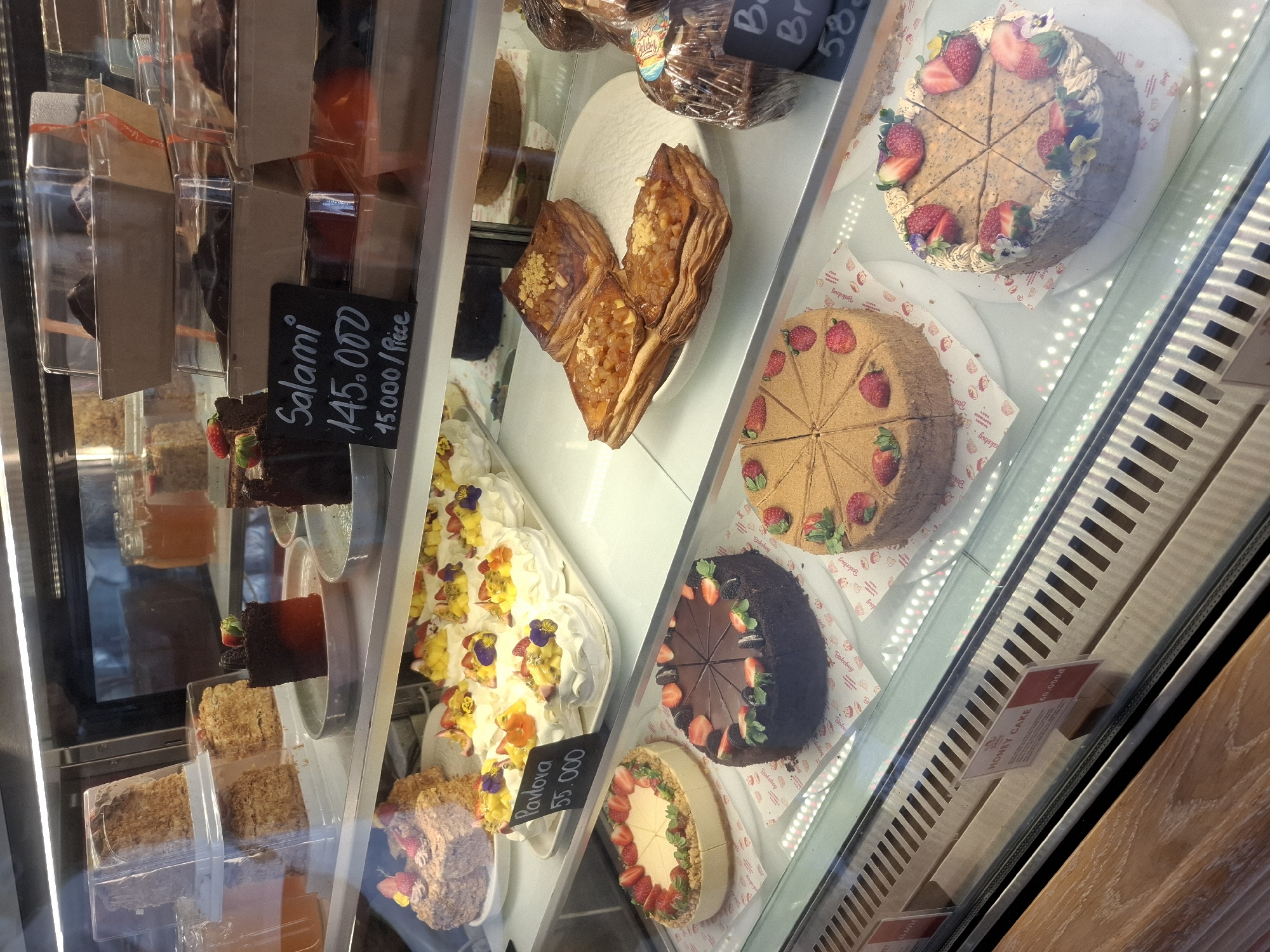 This is a medium shot, eye-level photograph of a bakery display case filled with various cakes and pastries. The display case is illuminated by overhead lighting, creating reflections on the glass. The background is dark, suggesting an indoor setting.
On the top shelf, several tall, rectangular cakes, possibly Napoleon cakes, are visible in clear plastic packaging. To their right, on a smaller shelf, are some jars of amber-colored liquid, possibly honey or syrup, and some wrapped desserts. In the center of the top shelf, there are individual slices of dark chocolate cake and a sign that reads "Salami 145.000 15.000/Piece".
The middle shelf features a large, pink cake decorated with what appear to be flowers and strawberries. To its right is a tray of meringue-based desserts, Pavlovas, topped with fruits like strawberries, passionfruit, and edible flowers, with a sign that reads "Pavlova 55.000".
The bottom shelf showcases several whole cakes and pastries. On the left, there is a round cake with a creamy base, decorated with strawberries and green leaves. Next to it is a dark chocolate cake, sliced into wedges and topped with strawberries and what look like blackberries. Further to the right, partially visible, are layered pastries with a fruit filling. On the far right, wrapped in plastic, are some dark, round desserts. A small blackboard next to these indicates "Br... 58...", likely prices.
The lower part of the image shows the interior of the bakery, with wooden shelving and metallic grates on the display unit. A label that reads "HONEY CAKE 60.000đ" is visible on the front of the display. The overall impression is of a well-stocked and appealing dessert selection.
This bakery is likely located in Nha Trang, Vietnam, given the currency symbol 'đ' and the style of the desserts commonly found in Vietnamese bakeries.