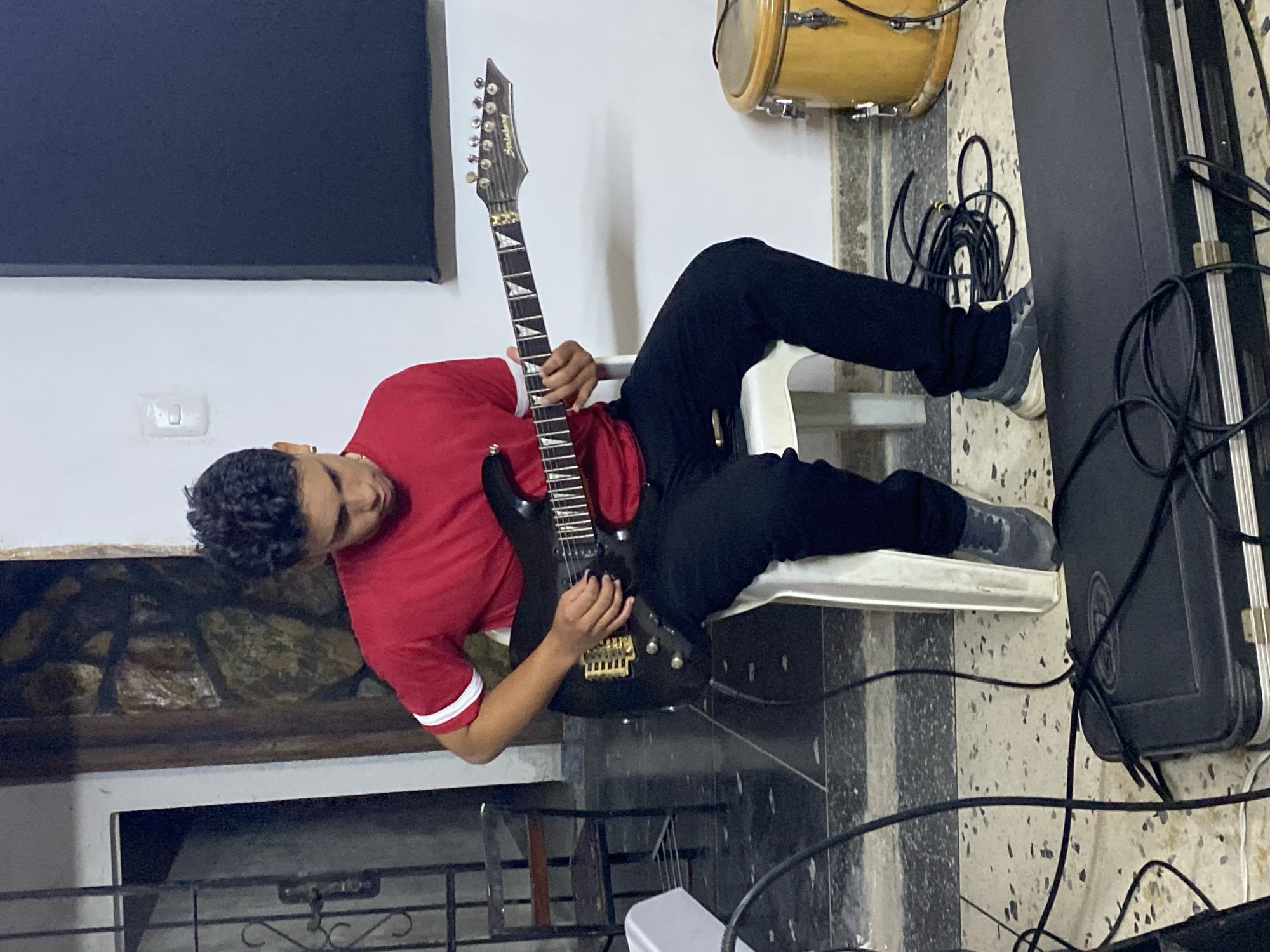 A young male individual is seated on a white plastic chair, focused on playing a black six-string electric guitar, identified by the "Steinberg" brand on its headstock and gold-colored bridge hardware. He wears a red short-sleeved t-shirt with white stripes on the sleeves, black pants, and grey sneakers. His left hand is positioned on the fretboard, while his right hand manipulates the strings near the bridge. The scene is an indoor environment, possibly a music practice space in Tinaquillo, Venezuela.
The floor is tiled with light-colored, speckled tiles. Numerous black audio cables are coiled and strewn across the floor, some resting on a black hardshell guitar case situated in the foreground. To the left of the individual, a dark metal gate or door with decorative scrollwork is visible, beyond which is a dark interior. A wall section adjacent to the gate features a textured, possibly stone or brick, finish. To the right, a white wall contains a rectangular black acoustic panel. A light switch or electrical outlet is present on the white wall. A light-brown percussion instrument, resembling a conga drum, is partially visible on the floor to the far right.