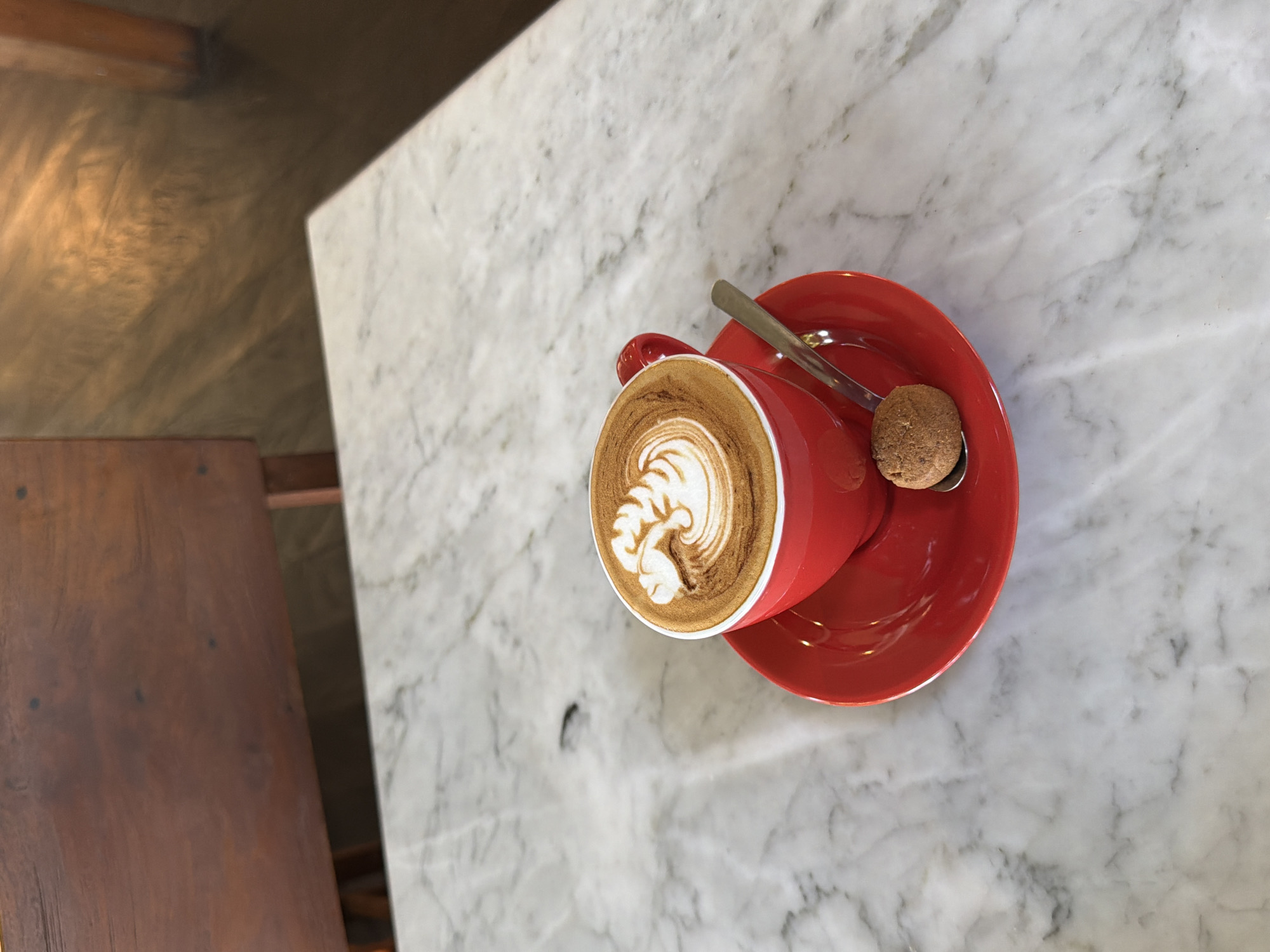 Overhead shot of a red cup of cappuccino with latte art, a red saucer, a cookie, and a spoon on a marble table in a cafe in North Kuta, Indonesia. A wooden table is visible to the left.