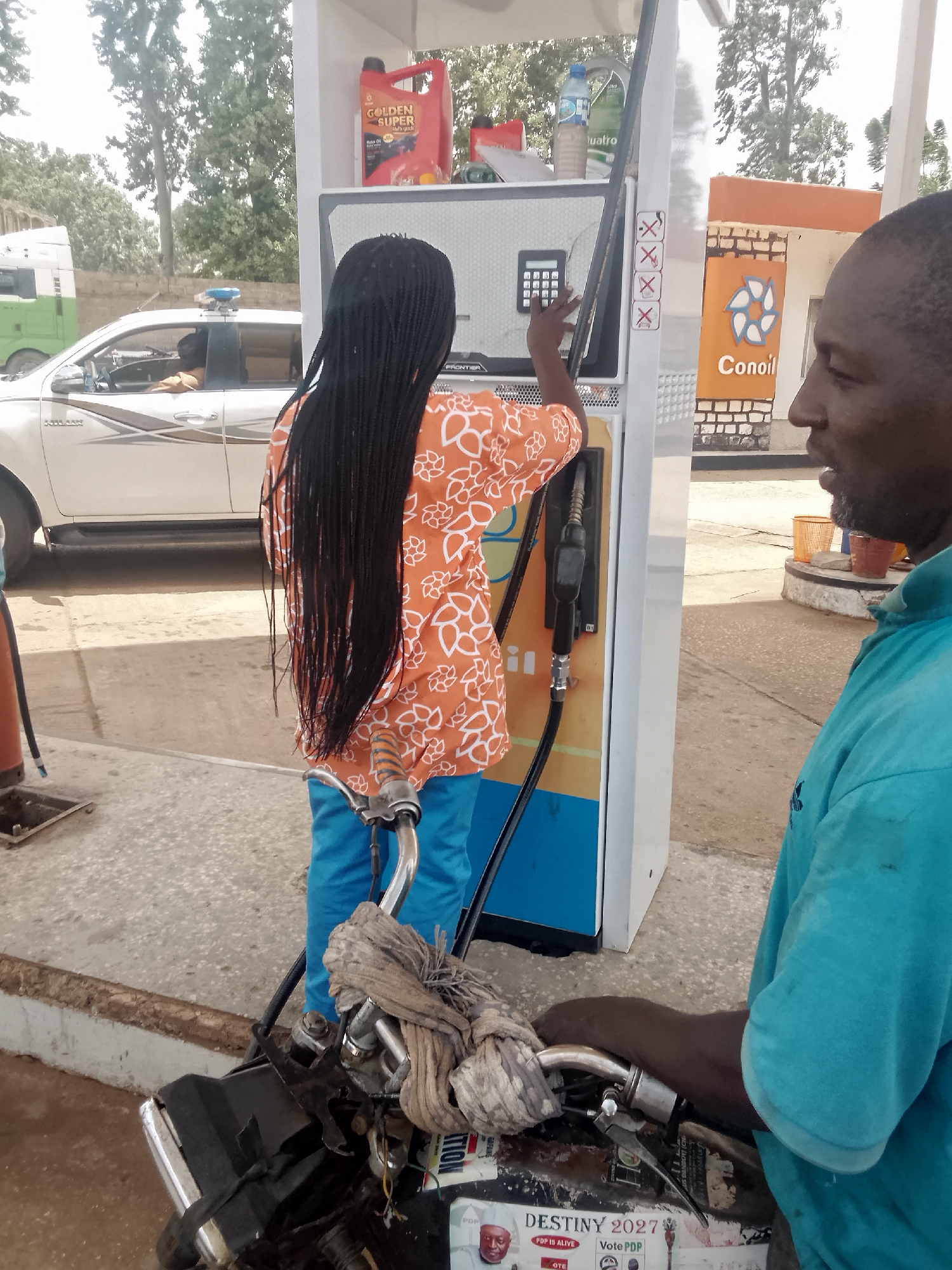 The image captures a scene at a Conoil gas station in Bukuru, Nigeria. A young person with long braided hair, wearing an orange floral shirt and blue pants, stands at a gas pump, appearing to enter information on the keypad. A man in a blue shirt watches, partially visible on the right.
In the foreground, a motorcycle is parked at the pump, its handlebars wrapped with a cloth. Stickers are visible on the front of the motorcycle, including one that reads "DESTINY 2027" and another with the phrase "Vote PDP."
In the background, a white pickup truck with a blue police light on top is parked. Beyond the truck, trees and a building are visible. Motor oil and water bottles are placed on top of the gas pump unit.