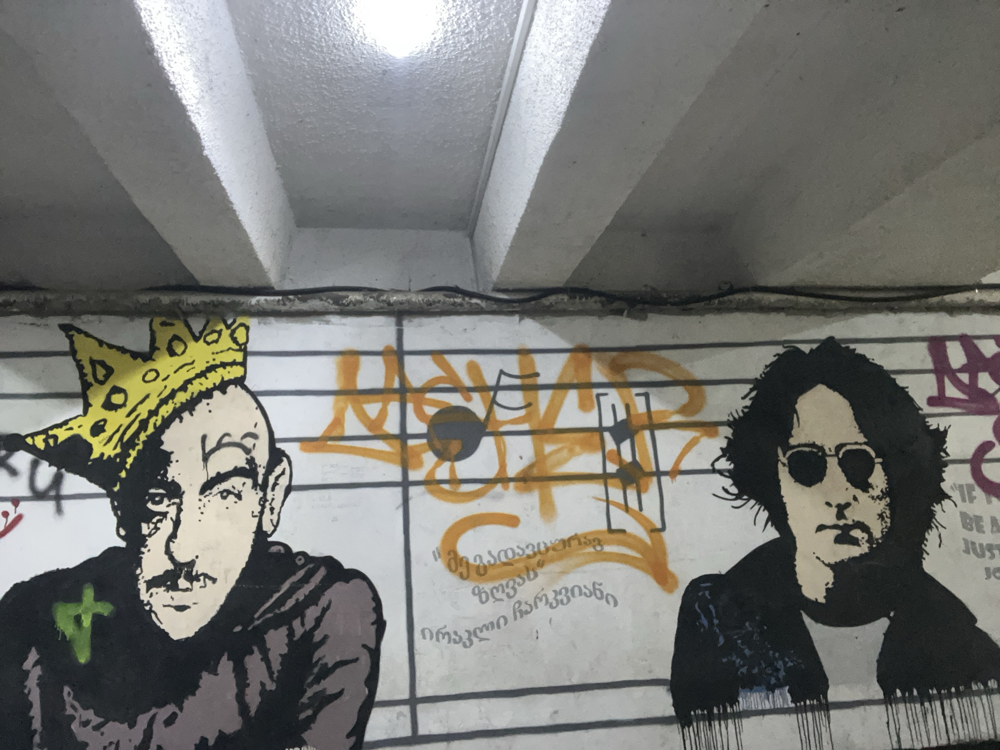 The image shows a section of graffiti art on a tiled wall, likely in an underpass or tunnel. The art depicts two portrait-style figures. On the left is a bald man with a mustache wearing a crown. A green mark resembling a cross adorns his chest. To the right is a figure resembling John Lennon, depicted with dark hair, sunglasses, and a jacket. Between them is spray-painted graffiti in Georgian script and a musical staff with notes. The setting appears to be a public area, and the condition of the wall indicates it is subject to weather exposure. The location is likely in Tbilisi, Georgia.