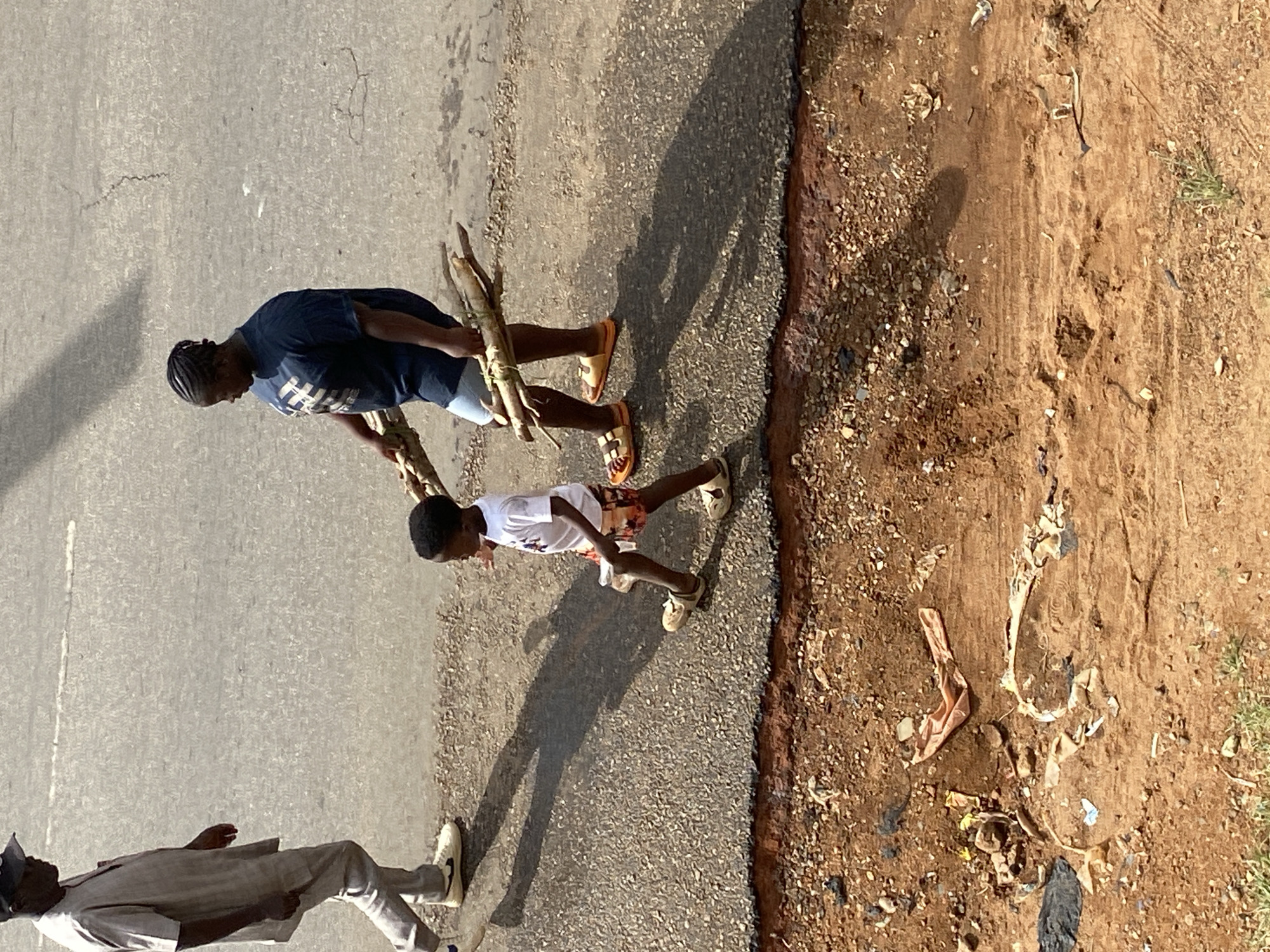The image shows three people walking along a road in Jos, Nigeria. One person is carrying a bundle of sticks. The road has a paved surface on one side and a dirt shoulder on the other, with trash scattered on the dirt. The lighting suggests it is daytime.