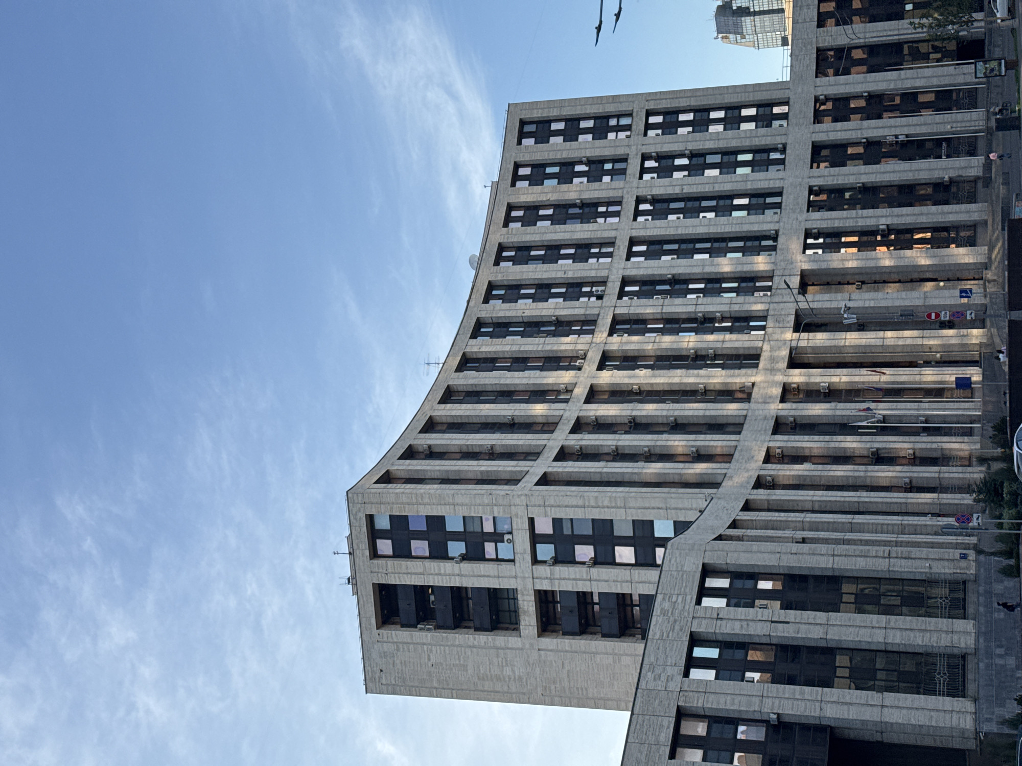 The image shows a modern, curved office building in Moscow, Russia. The building is clad in light-colored stone and features a facade with numerous vertical elements and dark, rectangular windows. The windows reflect the blue sky and some clouds. There are balconies on the upper floors of the building. Several air conditioning units are visible mounted on the exterior walls. In the foreground, a lower section of a building with similar architectural style is visible, with a dark glass facade. Street signs and a few people are present near the entrance of the building. The sky is clear and blue with some wispy clouds. In the background, another tall building with glass windows can be seen. The overall impression is of a contemporary urban landscape.