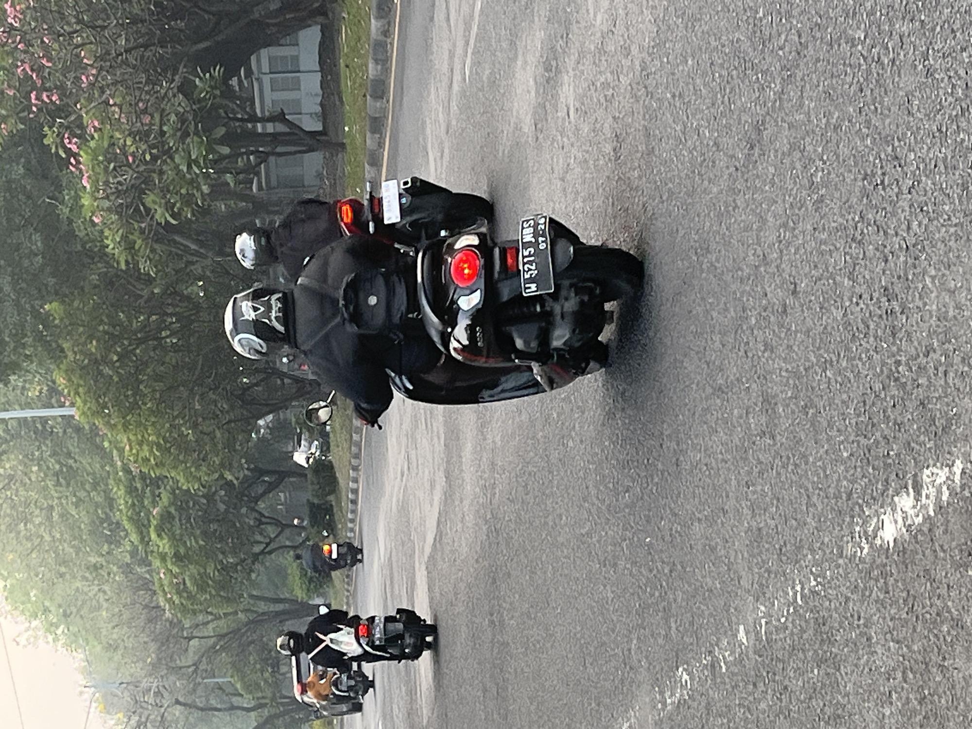 The image depicts a road scene in Sumorame, Indonesia, with several individuals riding motorcycles. The primary focus is on a person in the foreground, riding a black scooter with the license plate "W 5215 NBS 07-26". This rider is wearing a black jacket and a helmet with a white and black design. Another motorcycle, also black, is in front of this rider, and its rider is wearing a black jacket and helmet. Further ahead on the road, a third motorcycle, appearing to be red or dark colored, is visible with its rider wearing a helmet and dark clothing. In the background, on the left side, two more motorcycles and their riders are partially visible, one with a person wearing an orange jacket. The road is paved and has white lane markings. Lush green trees line the right side of the road, with some flowering trees displaying pink blossoms. The sky appears overcast or hazy. The overall scene suggests a typical day of motorcycle transportation in this Indonesian locale.