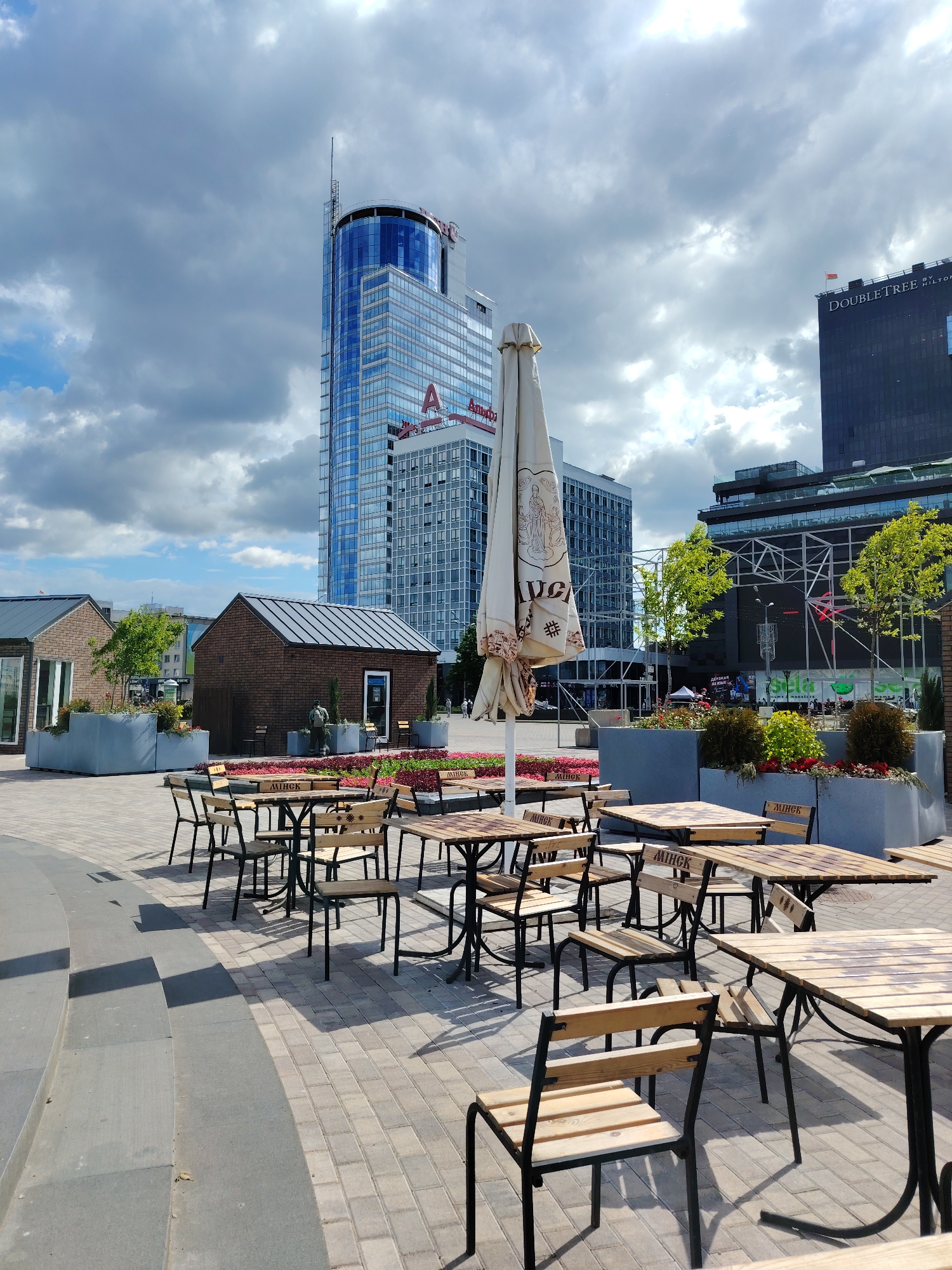 This outdoor daytime photograph captures a public square or patio area in Minsk, Belarus, featuring numerous empty tables and chairs arranged for outdoor dining. The ground is paved with light-colored bricks, and a curved concrete seating area is visible in the foreground. Several planters filled with vibrant red, pink, and yellow flowers, along with small trees, add greenery to the scene.
In the mid-ground, a large, folded beige outdoor parasol stands prominently, displaying what appears to be a brewery logo with a female figure and the text "АЛIВАРЫЯ" (Alivaria), a known Belarusian brand. To the left, two small, modern brick-clad buildings with dark roofs are situated, one featuring a large window and the other a door, possibly kiosks or small shops.
The background is dominated by modern high-rise buildings. A tall, glass skyscraper with a rounded blue facade stands out, bearing a red "A" logo and the text "АМАТАР" (AMATAR) on its side. To the right, another dark-colored building clearly displays "DOUBLETREE by Hilton" on its facade, with a small flag, possibly the flag of Belarus, visible on its roof. Another building in the background also shows "АМАТАР". The backs of some chairs, tables, and planters have the word "MINCK" or "MINSK" inscribed, confirming the city.
The sky is partly cloudy, suggesting bright daylight with diffuse lighting, and shadows indicate it's mid-day. The area appears quiet and unoccupied, with no significant activity taking place, giving the impression of a space ready for use or currently closed.