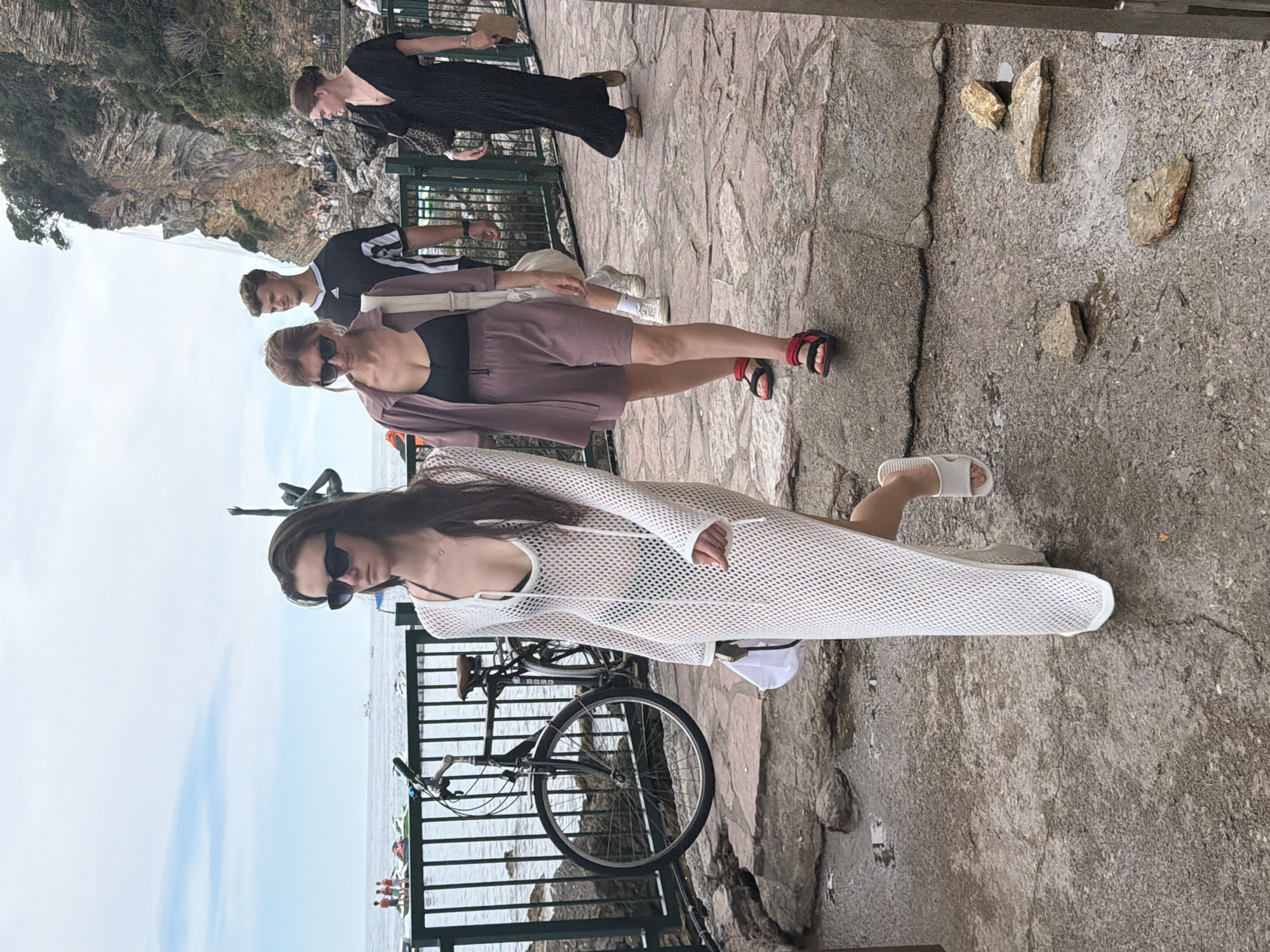 The image captures a view of the Budva Riviera in Montenegro, specifically along a pedestrian walkway near the coast. In the foreground, a young woman with long dark hair and sunglasses walks towards the viewer, wearing a white crocheted dress over a black bikini and white slides. Behind her, a woman with reddish-brown hair and sunglasses walks in a mauve colored coordinated set, wearing red sandals. A young man in an Adidas t-shirt follows them. Further in the background, another woman in a black dress walks away from the viewer. A bicycle is parked to the left, secured by a dark green metal fence. The walkway is made of textured stone, with scattered rocks and some wet patches. Beyond the fence, the Adriatic Sea is visible with a few boats and people engaged in water activities. A rocky cliff face, covered in greenery, rises dramatically on the right side of the image, with a waterfall cascading down its side. The sky is cloudy.