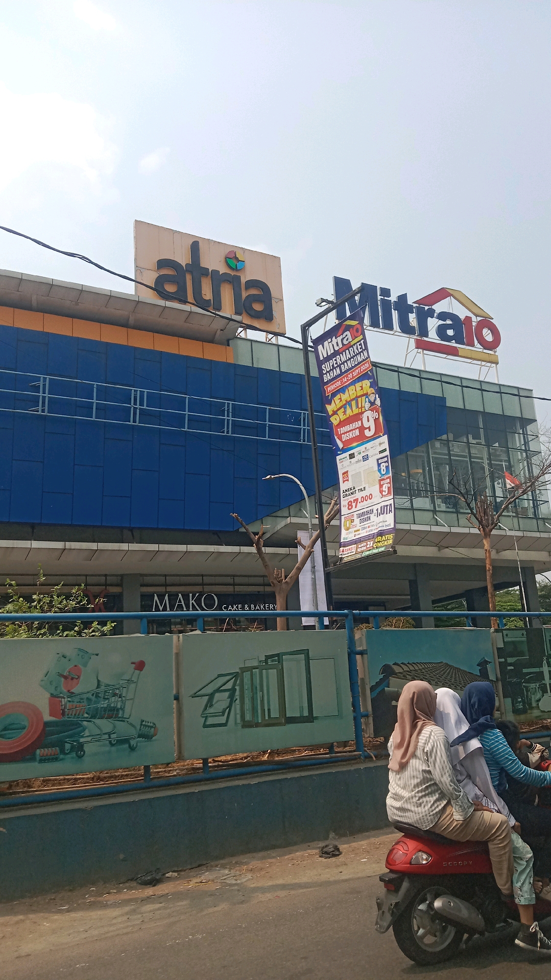 The image captures a street view in Bekasi, Indonesia, focusing on a large retail establishment. Dominating the scene are the prominent logos of "Atria" and "Mitra 10," indicating these are the names of the stores. "Atria" appears to be a furniture store, while "Mitra 10" is identified as a supermarket for building materials. A large banner for "Mitra 10" is visible, advertising a "Member Deal" with a 9% discount and other promotions like "Tambahan Diskon" (additional discount) and "Gratis Ongkir" (free shipping).
Below the main store fascia, the sign for "MAKO CAKE & BAKERY" is clearly displayed, suggesting a smaller establishment within or adjacent to the larger complex. The building itself has a modern architectural design with a blue facade and orange accents.
In the foreground, a red scooter is carrying three people, two women wearing hijabs and another person partially obscured. They are moving away from the camera, suggesting a busy street environment. The background shows a clear sky with some clouds, typical of the tropical climate. A fence with graphics depicting various building materials like windows and a shopping cart adds to the commercial context of the location. The overall scene depicts a commercial district in Bekasi, with a focus on retail shopping and daily life activities.