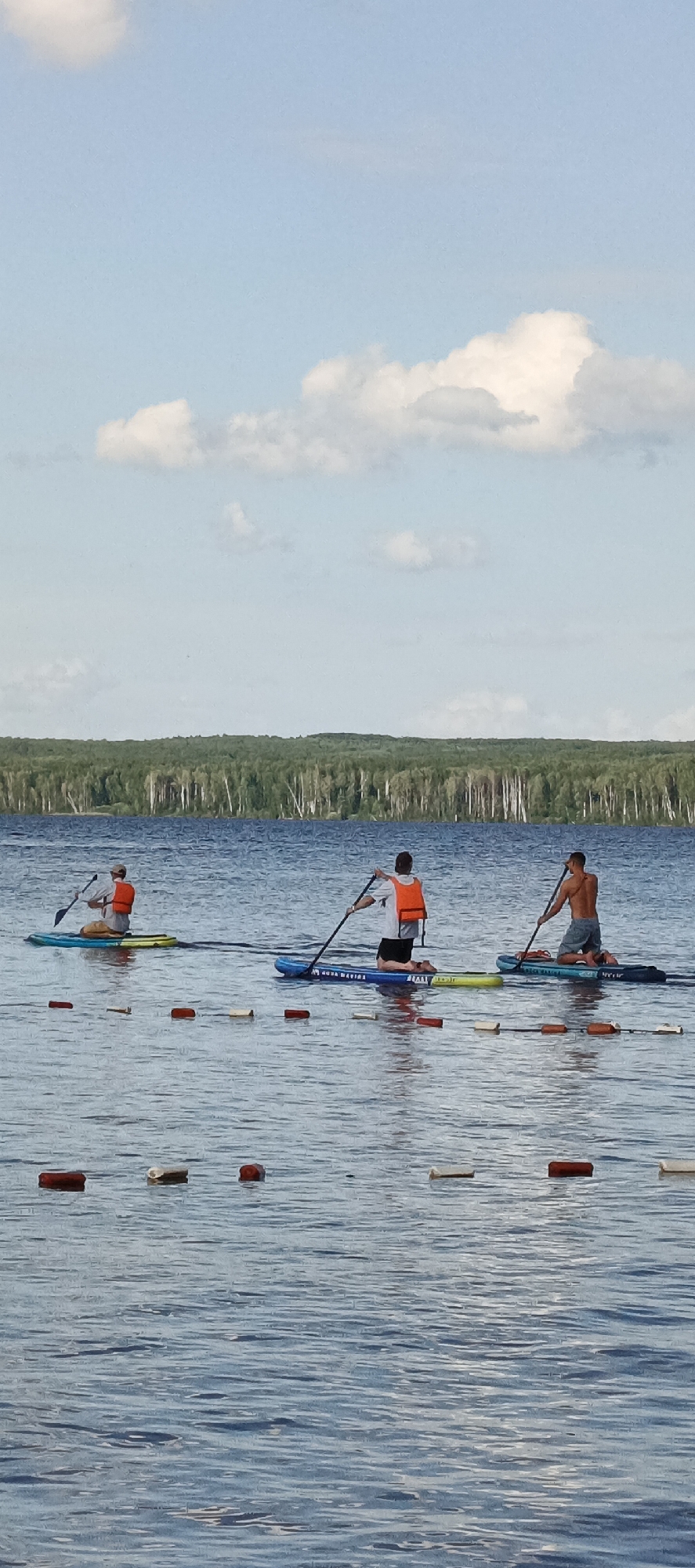 Three people are paddleboarding on a lake with a forest in the background. The sky is blue with scattered white clouds. The water is calm and reflects the sky. There are buoys in the water in front of the paddleboarders. The people are wearing orange life vests and appear to be enjoying the activity. The scene suggests a warm, sunny day. The location is Chaikovsky, Russia. No visible text is present in the image.