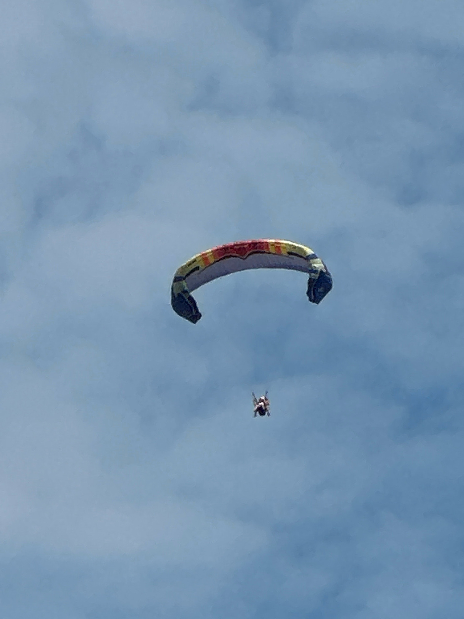 A paraglider is pictured in the sky above Boreti, Montenegro, against a backdrop of a blue sky with scattered clouds. The paraglider, a large, crescent-shaped canopy, is multi-colored with stripes of yellow, red, and blue. Beneath the paraglider, a person is suspended, appearing to be in the process of paragliding. The image captures a moment of flight and recreational activity.