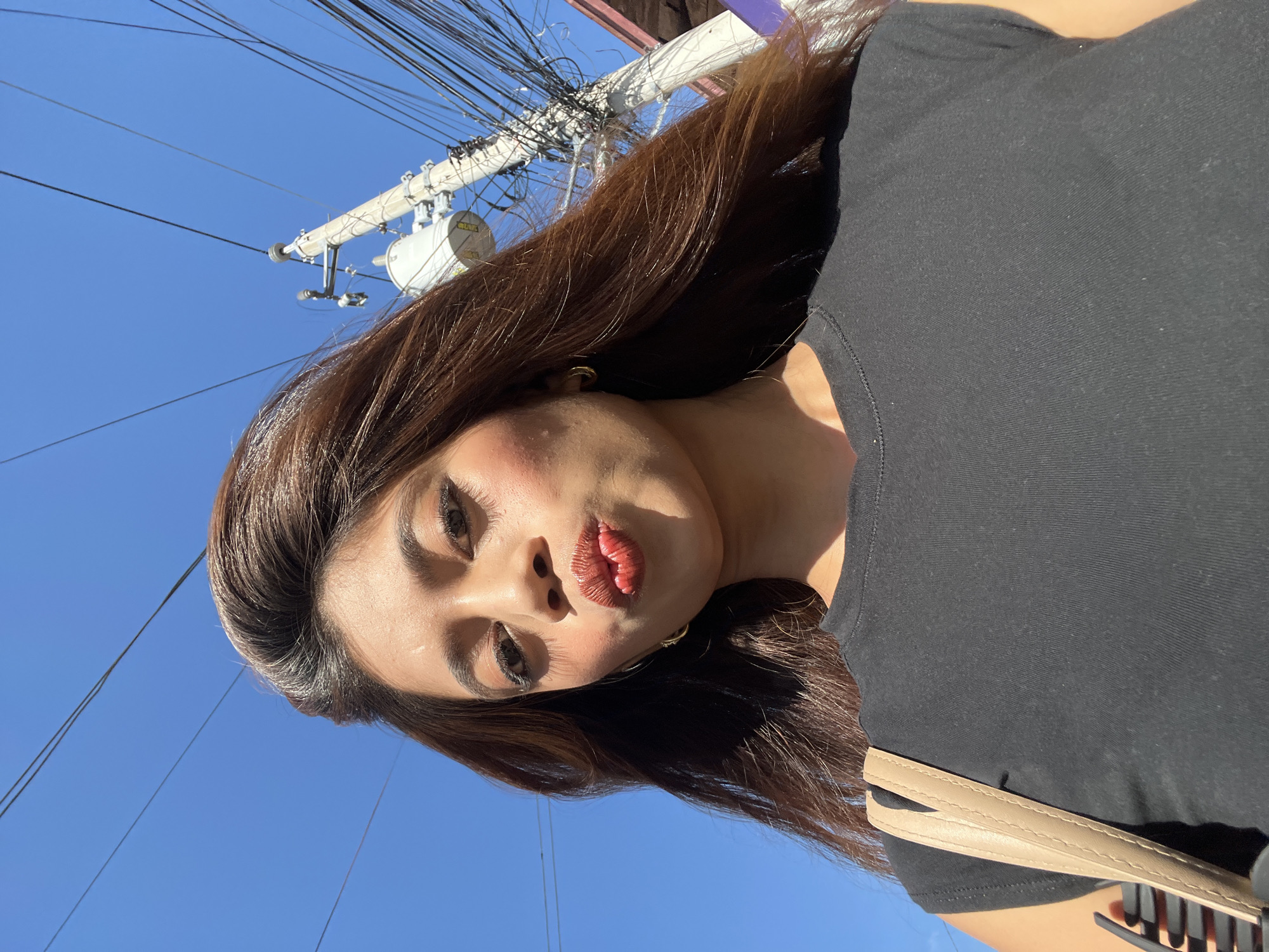 A low-angle, medium shot captures a woman with long, brown hair, who is making a kissing gesture towards the camera. She is outdoors, with a clear blue sky overhead. A utility pole with numerous wires is visible in the background, to her right. The woman is wearing a black, short-sleeved t-shirt and a beige purse strap is visible over her left shoulder. The scene is indicative of an urban environment in Davao City, Philippines, suggested by the characteristic utility pole and wire configurations common in the region.