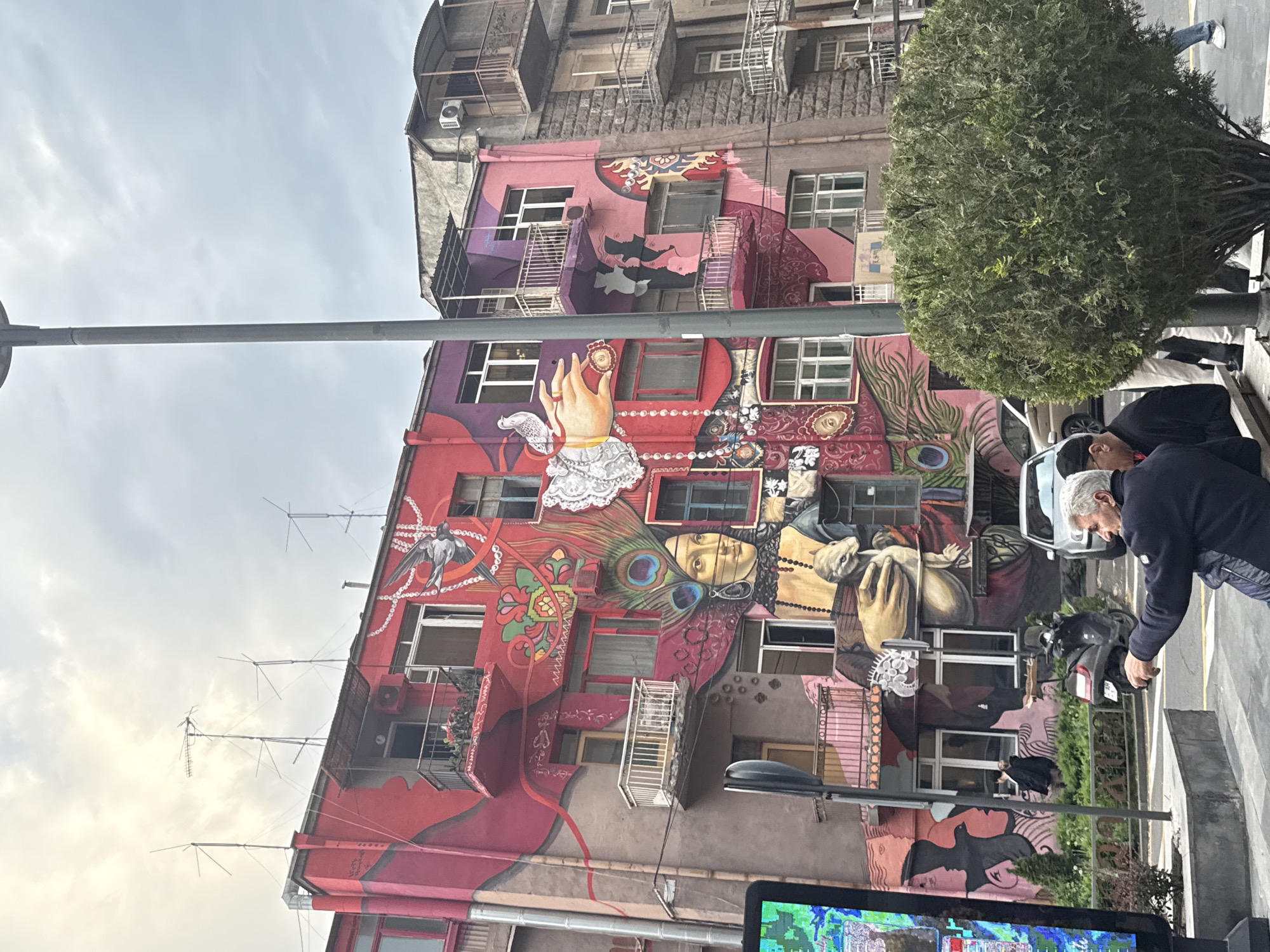 The image captures a building adorned with vibrant murals. The structure is multi-storied, with an older section on top. The murals feature a variety of images: hand with lace sleeve, faces, and peacock feathers. There are people and vehicles present in the foreground, although obscured. One person is interacting with a scooter, another person can be seen from the back. The overall setting appears to be an urban street scene.