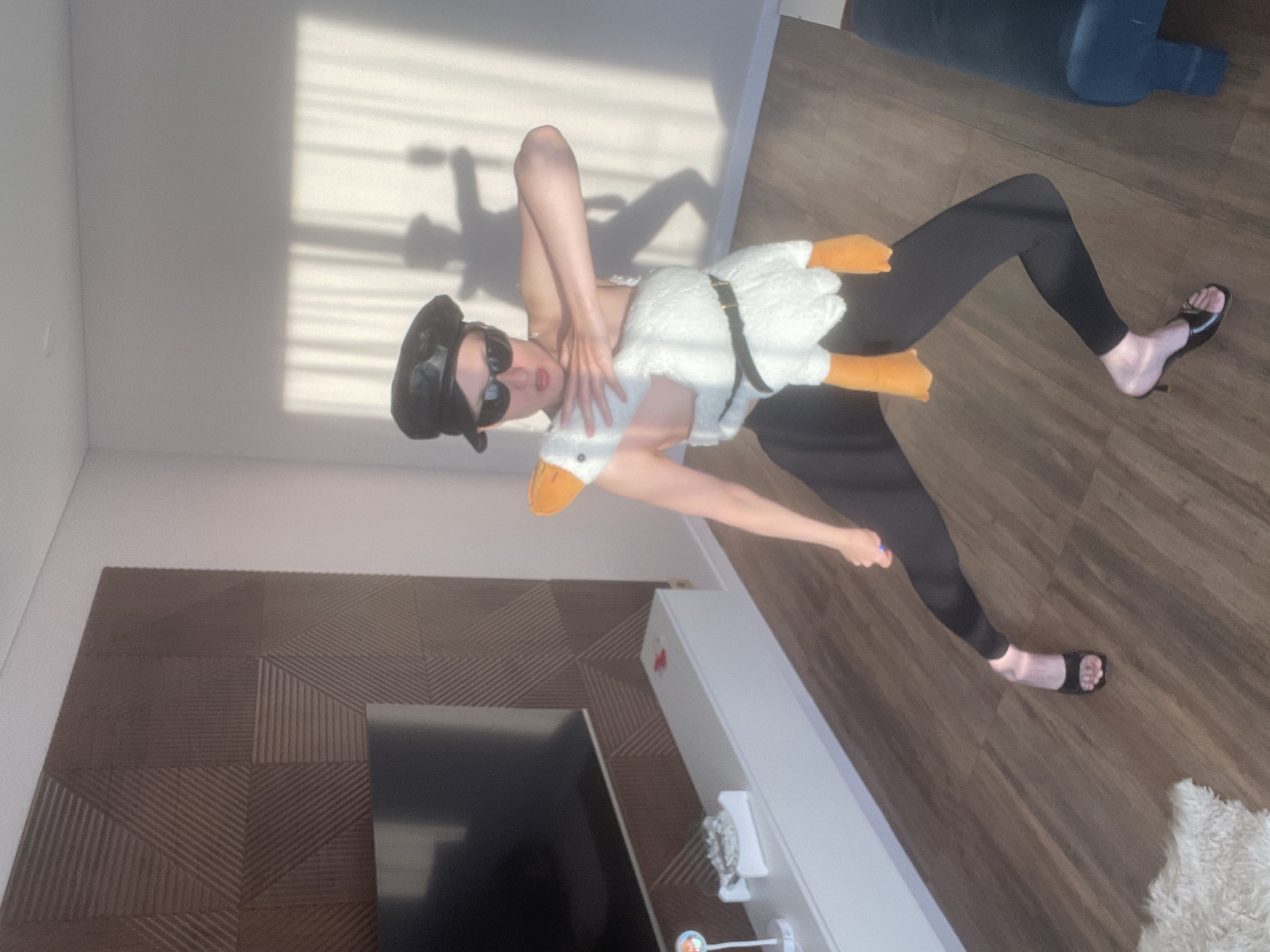 A person is captured in a playful pose indoors, dressed in a whimsical duck costume. The individual, appearing female, wears black leggings, black high-heeled sandals, dark sunglasses, and a black beret. They hold the white, plush duck costume, which features orange feet and a beak, around their torso, secured by a black belt. Their left arm is bent upwards while their right arm extends forward.
The setting is a modern room with light walls and a dark, possibly wooden, floor. Strong sunlight from a window casts distinct, horizontal shadows from window blinds onto the wall behind the person, indicating it is daytime, likely morning or late afternoon. Geometric patterns of textured wooden panels adorn a wall to the left, beneath which a large, dark television screen is partially visible. A white cabinet or shelf stands to the right. A dark blue upholstered piece of furniture, possibly a sofa or chair, can be seen in the upper right background. A fluffy, light-colored rug is also visible on the floor in the lower right. No text is visible in the image.