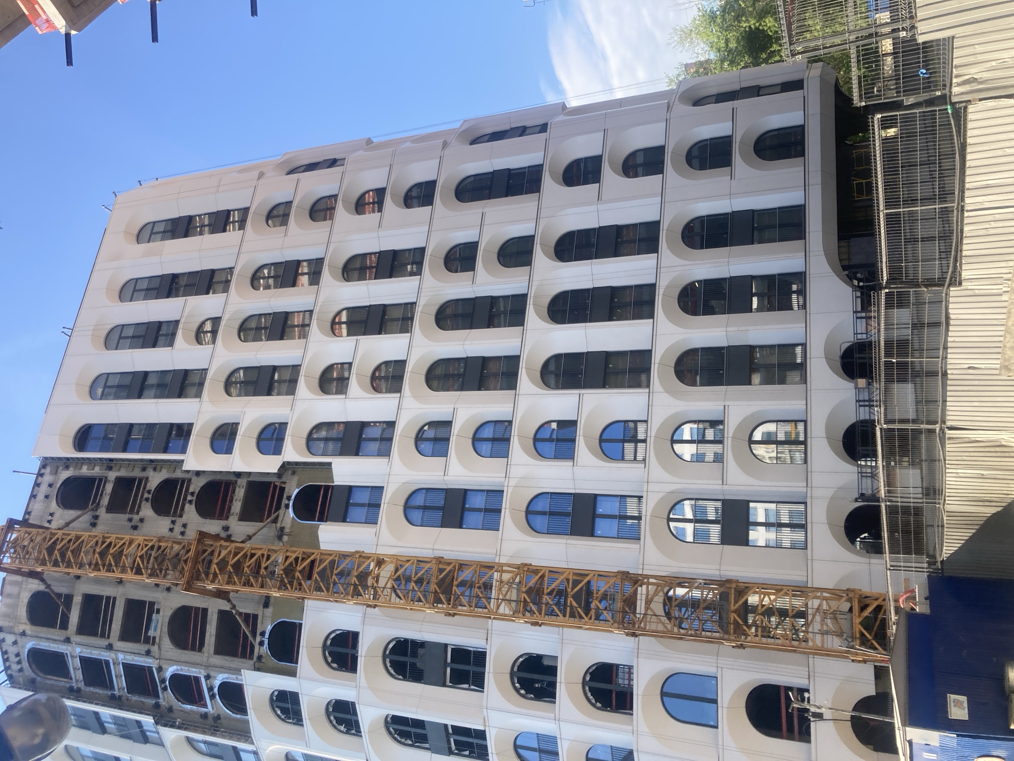 The image shows a high-rise building under construction in Moscow, Russia, with a yellow crane positioned against its facade. The building features a modern architectural design with repeating arched windows and balconies. The lower levels of the building are partially obscured by construction fencing and temporary structures. The sky is clear and blue, indicating a sunny day.