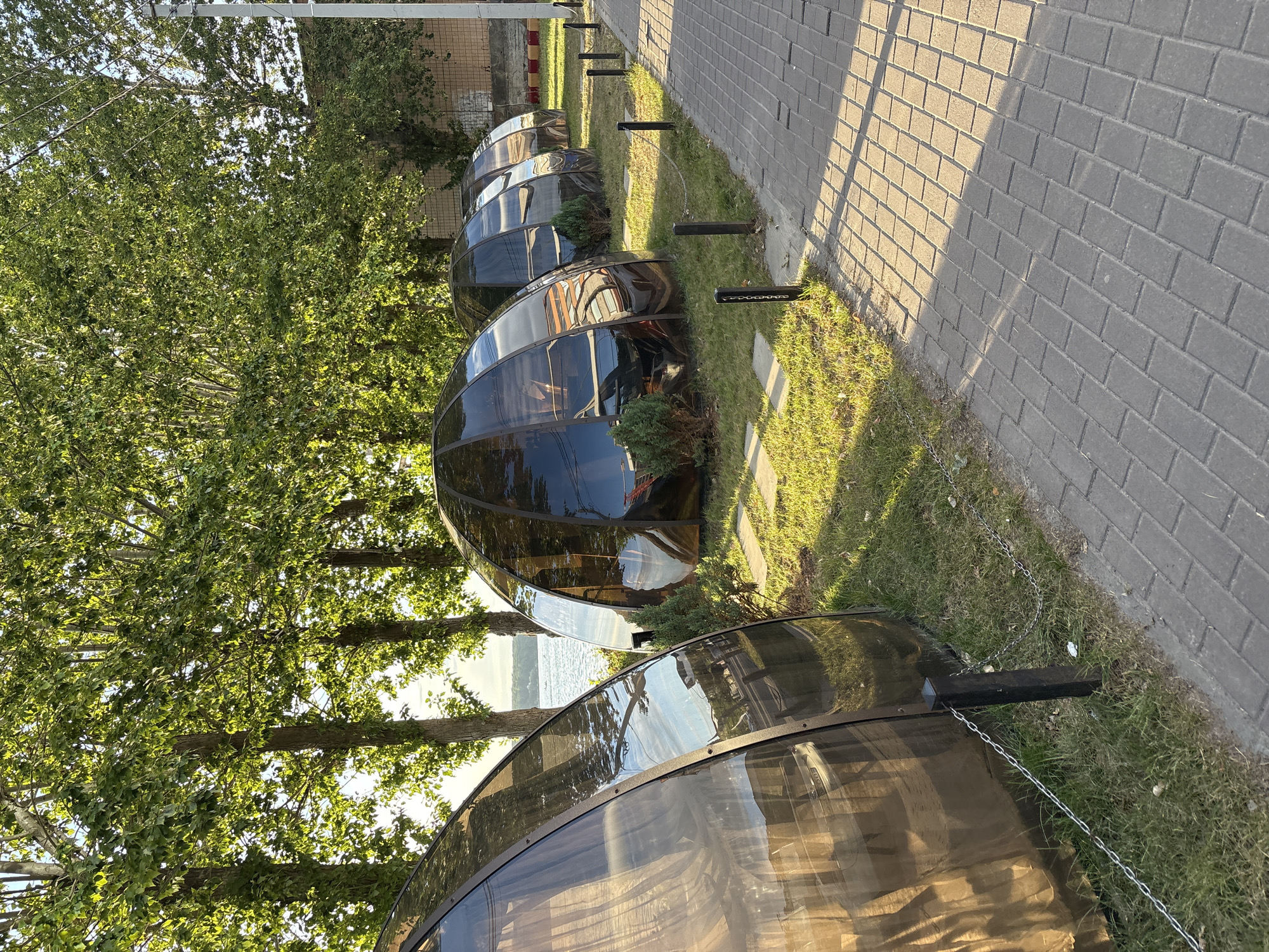 The image shows a row of transparent geodesic domes situated on a grassy area next to a paved walkway. These domes appear to be enclosed seating areas, possibly for a restaurant or cafe, with some furniture visible inside. In the background, a body of water, likely a lake or river, is visible, bordered by trees. The scene is bathed in the warm light of the sun, casting shadows on the walkway. The domes are dark, reflective, and spherical in shape, with metal framing. Small, manicured bushes are planted around the domes. A metal chain fence runs along the edge of the grass where it meets the walkway, supported by dark posts. In the far background, a brick building and utility pole can be seen. The overall setting suggests an outdoor dining or recreational area by the water. This location is identified as Ternopil Lake (Teremky), Ternopil, Ukraine.