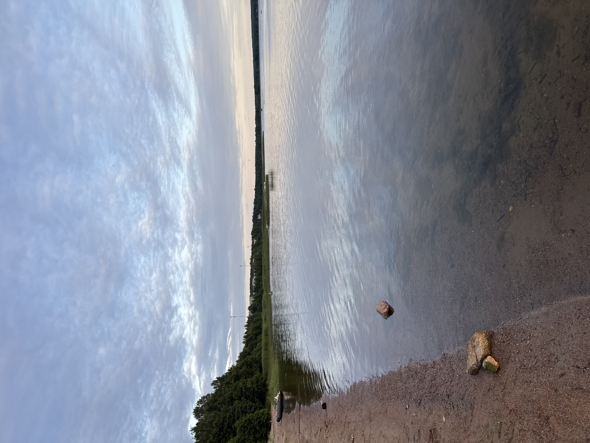 The image depicts a serene lake scene on the outskirts of Glebychevo, Russia. The water is calm, reflecting the overcast sky and the dense green treeline that borders the opposite shore. In the foreground, a sandy and pebbly beach is visible, with a few scattered rocks. An inflatable boat, dark gray in color, rests on the shore. The sky is filled with a layer of scattered, pale clouds. The overall impression is one of a tranquil, natural landscape.