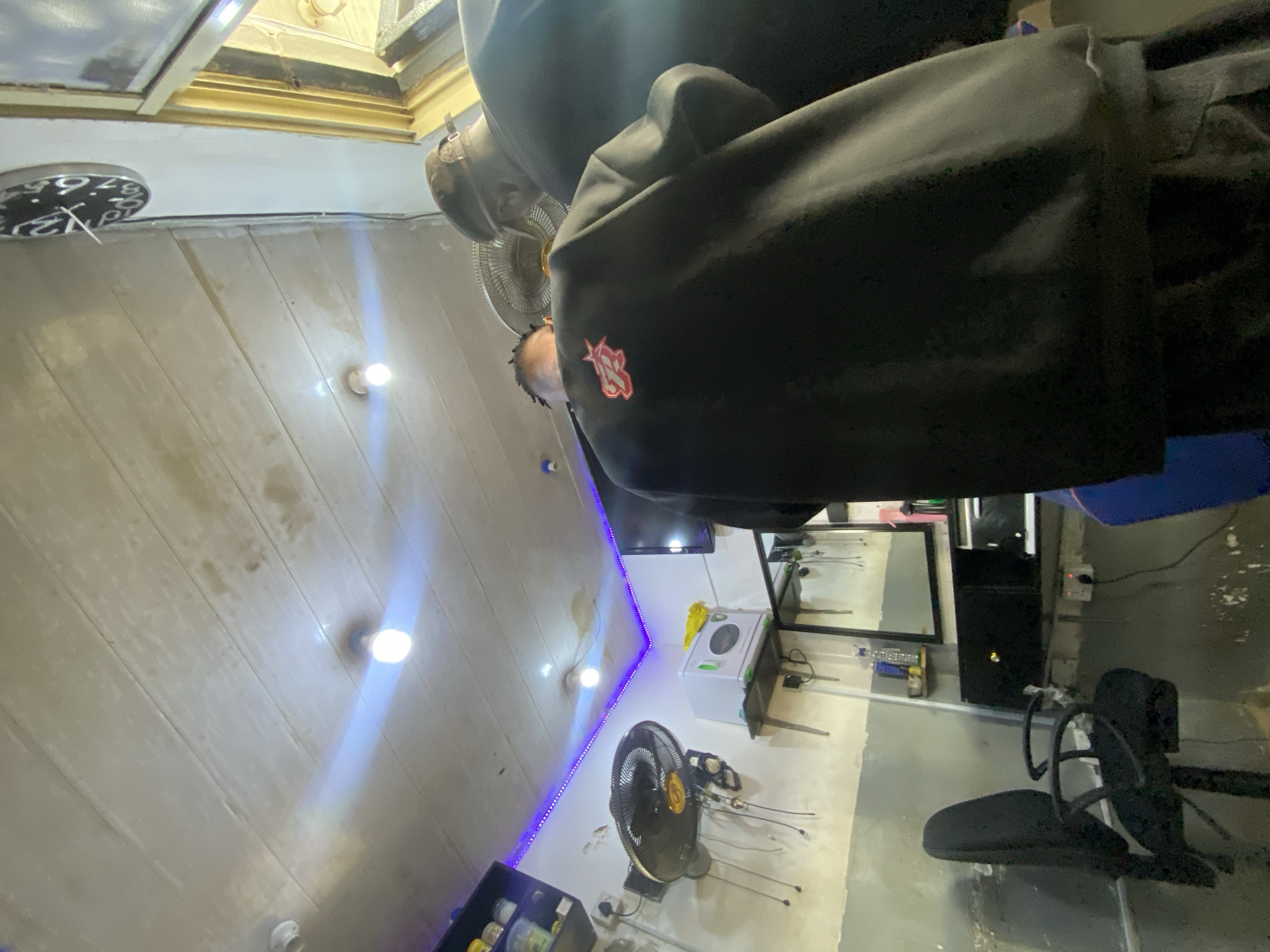 A photograph taken indoors in Ado, Nigeria, presents a slanted view of a barber shop setting. The perspective is skewed, showing a diagonal orientation of the room. Dominating the foreground is a person wearing a black hooded garment with a red logo visible on the left side. In the background, a seated individual appears to be receiving a haircut. The shop interior features white paneled walls illuminated by multiple evenly-spaced light bulbs. A wall-mounted fan with gold accents is situated near a small white appliance. A mirror and various barbering tools can be seen along the wall. A black barber chair is present in the scene. A television is mounted on the wall. A decorative blue LED strip runs along the wall.