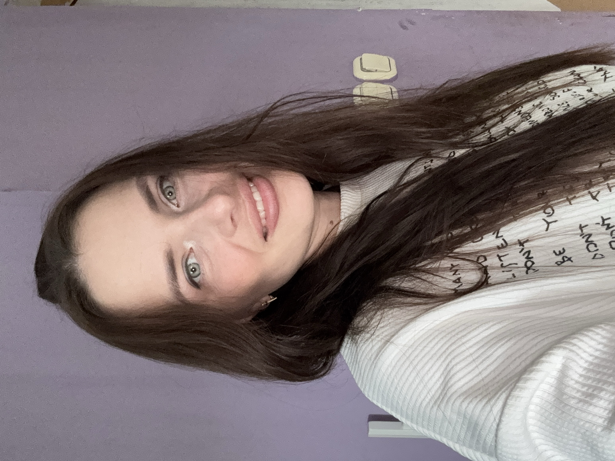 The media file shows a close-up, vertically oriented selfie of a young woman with long, dark brown hair. She is looking directly at the camera with a slight smile, revealing white teeth and pink lips. Her eyes are a striking blue-green. She is wearing a white, ribbed sweater with black text printed on it. Some of the visible text includes "DON'T", "LISTEN", "WANT", and "BE". The background is a plain, light purple wall with two white rectangular objects mounted on it. The lighting suggests it is daytime, and the indoor setting appears to be a bedroom or personal space. The woman's expression conveys a relaxed and happy mood. No specific weather or event is indicated. The implied location is Rusalevka, Russia, based on the provided context.