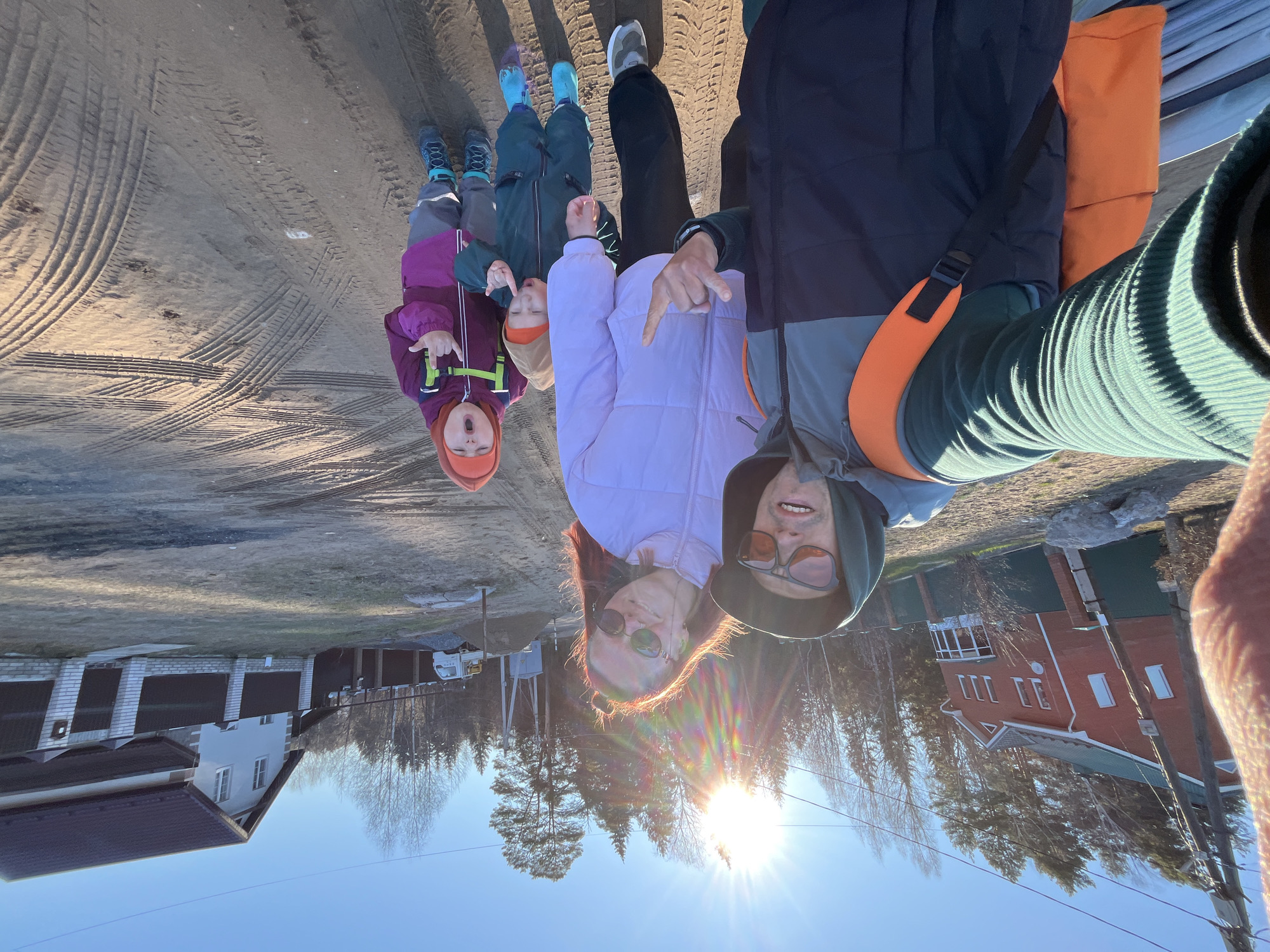 An outdoor, eye-level selfie captures a family of five in Chaikovsky, Russia, on a bright, sunny day. The image, oriented sideways, shows two adults and three children on a dirt ground covered with numerous tire tracks. The sun is low in the sky, casting a strong flare and suggesting either early morning or late afternoon.
In the foreground, a woman with reddish-brown hair and sunglasses, wearing a light purple jacket, smiles towards the camera. Beside her, a man in a dark green hoodie, a jacket with grey, navy, and orange sections, and sunglasses, also looks at the camera. Both adults, along with one child, are playfully pointing downwards with their index fingers. The man has an orange strap from a bag or backpack across his shoulder.
Three children are visible behind the adults. One child wears a dark purple jacket, an orange hat, and green backpack straps, making an expressive face. Another child wears a dark green jacket and an orange hat. A third child is partially visible further back, wearing blue shoes and grey pants.
The background features several residential buildings and trees. To the left, a light-colored house with a dark roof, white windows, and brick pillars is visible. In the distance, a red brick house with a green roof can be seen through bare trees, indicative of late autumn, winter, or early spring. A white truck, possibly a utility or construction vehicle, is parked near one of the houses. The sky is clear blue. No discernible text is visible in the image.