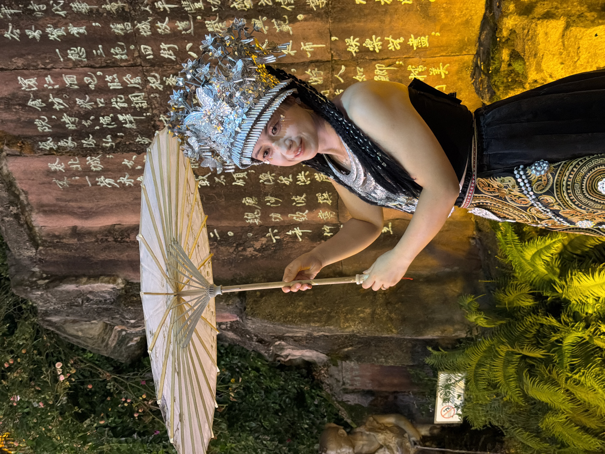 A person, adorned in elaborate traditional Chinese attire and a silver headdress, holds a white paper umbrella. The individual has dark braided hair and makeup that includes sparkling accents on their cheeks and eyelids. They are looking towards the viewer with a gentle smile. The background features a textured wall with glowing Chinese calligraphy projected onto it, giving the scene an ethereal and artistic feel. To the right of the person, lush green ferns are visible, illuminated by warm, yellow lighting that also casts a golden glow on the calligraphy. The overall setting appears to be an outdoor space at night, possibly a cultural performance or a decorated historical site. The calligraphy on the wall is in white and appears to be ancient script, adding to the cultural richness of the scene. The person's clothing is detailed, featuring intricate embroidery with gold and red accents, and a beaded necklace. The umbrella is made of delicate paper and bamboo spokes.