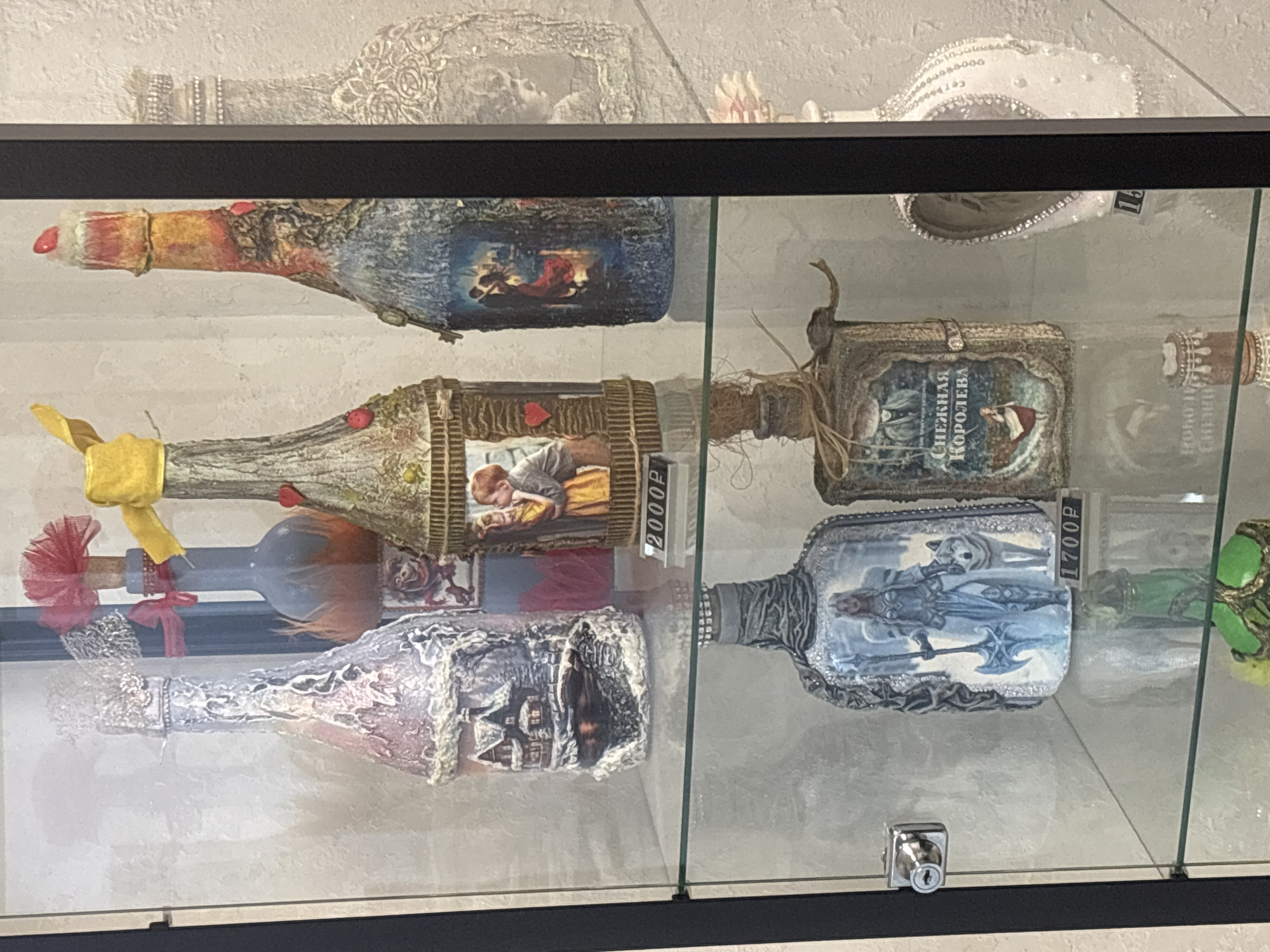 A glass-enclosed display cabinet, likely in a shop or exhibition space in Dolgoprudny, Russia, showcases several ornate, decorated bottles. The bottles are placed on glass shelves.
In the upper section of the cabinet, from left to right, are four bottles. The first bottle is decorated with a textured, silvery-pink material and features a miniature winter scene with a house. Next to it is a blue bottle adorned with red and orange feathery accents and a red bow. The third bottle has a textured, brown and gold exterior and incorporates a framed picture of a couple kissing. The fourth bottle has a textured surface with blue and orange hues and depicts a couple dancing. A price tag reading "2000P" is visible near the third bottle.
The lower section of the cabinet displays three more bottles. The first on the left is decorated with a silver-toned pattern and features an image of a woman in armor with an axe, accompanied by a wolf. A price tag reading "1700P" is visible below this bottle. Next to it is a bottle with a book-like decoration, featuring an image of a woman and the text "Снежная Королева" (Snow Queen). To the far right is a white bottle embellished with pearls and rhinestones, with a price tag reading "15" partially visible.
Other decorative elements and reflections are present within the cabinet and on the glass. The background appears to be a textured wall. A silver lock mechanism is visible on the left side of the cabinet.