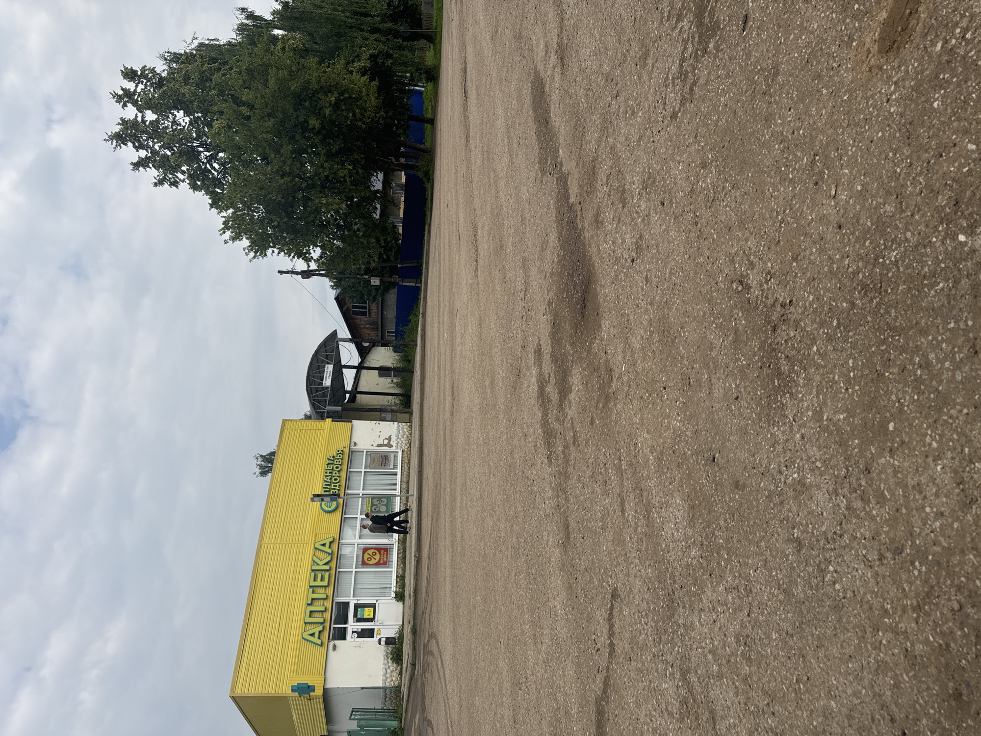 In Chastyye, Russia, a low-angle shot captures the exterior of a bright yellow building with "АПТЕКА" (Pharmacy) and "ПЛАНЕТА ЗДОРОВЬЯ" (Planet of Health) signage in large green letters. The building has a white base with stone accents and large windows, some displaying signs with percentages and medical-related imagery. Two individuals, a man and a woman, are walking away from the building on a sidewalk in front of it. To the right of the pharmacy, a covered structure with a white roof and support beams is visible. Beyond the building, mature trees with dense green foliage are prominent. The foreground consists of a wide, uneven, light-brown paved area, possibly a parking lot or open space, with visible tire marks and darker patches indicating moisture or wear. The sky above is overcast with scattered white clouds. A street lamp and a dark blue fence can be seen in the middle ground behind the pharmacy and trees. The overall scene depicts a commercial area in a town.