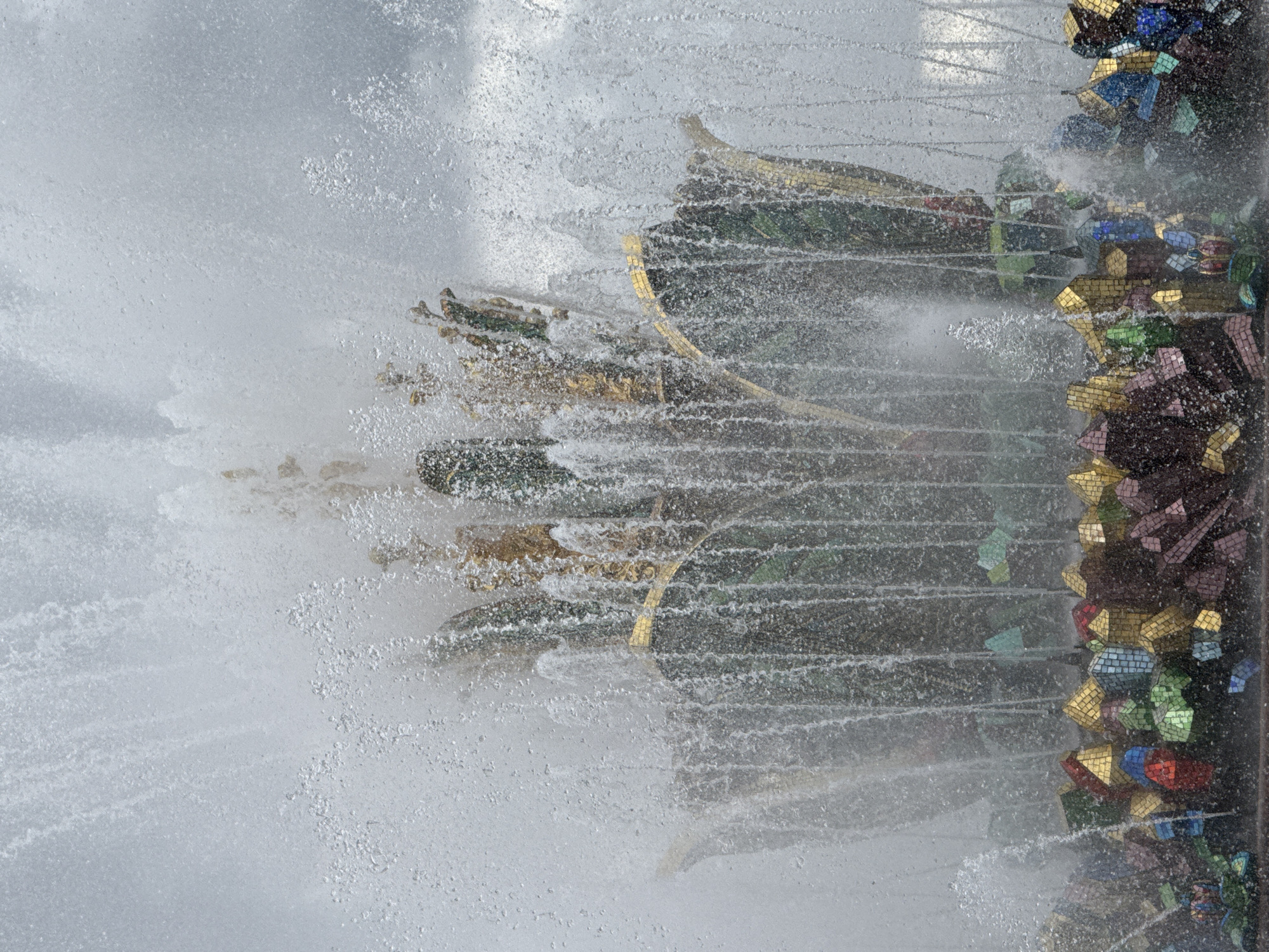 This is a close-up, eye-level, medium shot of the "Stone Flower" fountain, also known as the "Friendship of Peoples" fountain, located in VDNKh (Exhibition of Achievements of National Economy) in Moscow, Russia. The fountain is heavily active, with water spraying and cascading from its structure, obscuring some details. The central part of the fountain, resembling a large stylized flower, is adorned with gold and green mosaic tiles. The base of the fountain is decorated with a collection of colorful, angular mosaic pieces, appearing like clustered geometric forms in shades of red, blue, green, gold, and purple. The surrounding environment is filled with spray and mist from the water, creating a soft, diffused background, with a hint of a cloudy sky overhead. There are no people visible in the frame.