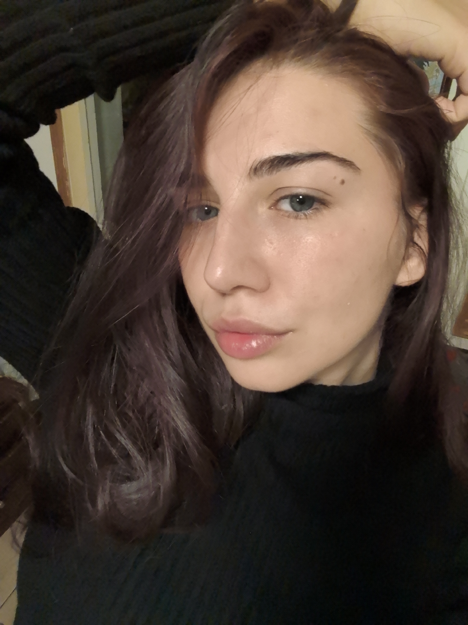 The scene is an indoor setting in Stara Zagora, Bulgaria. A young adult female is depicted in a close-up, chest-up portrait, gazing directly at the camera. She has long, dark brown hair with reddish-purple undertones, styled with a side part, falling across her shoulders and partially framing her face. Her eyes are light blue-grey, and her eyebrows are dark and well-defined. A small, dark mole is visible above her left eyebrow, and another lighter spot is present below her right eye. Her skin exhibits a natural, slightly dewy appearance. Her lips are full, pink, and have a glossy finish. She is wearing a black, ribbed turtleneck garment. Her left hand is partially visible in the upper left quadrant, near her head, and her right hand is seen in the upper right, appearing to adjust or hold her hair. The background is out of focus, showing indistinct dark and light beige or yellow tones, consistent with an interior wall or door frame.