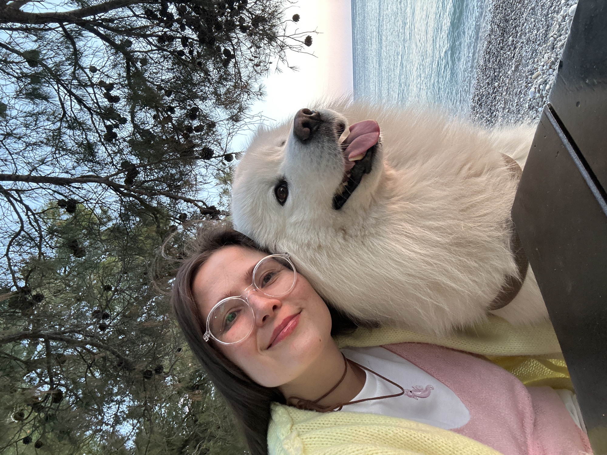 A woman in a yellow and pink cardigan, white shirt, brown choker, and clear-framed glasses is posing for a selfie with a large white Samoyed dog, close to the beach in Šušanj, Montenegro. The dog has its tongue out. The background includes a pine tree with pine cones, the Adriatic Sea, and a pebbled beach.