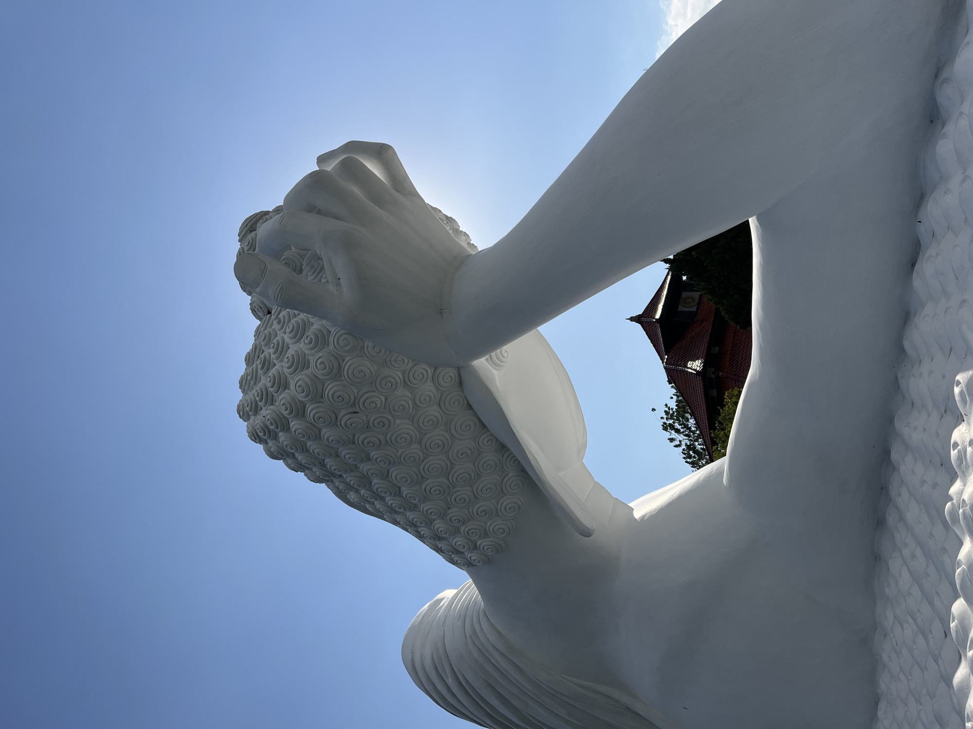 A low-angle, close-up, outdoor, eye-level shot showcases a large, white statue of a reclining Buddha against a bright blue sky with a hint of the sun creating a slight glare. The sculpture is white and appears to be made of smooth, polished material. The Buddha's head is tilted back, resting on one hand which is positioned under its head. Its hair is rendered in intricate, swirling patterns. The body of the Buddha is partially visible, with a flowing robe draped over its shoulder. In the background, a traditional building with a red tiled roof is visible amongst green trees, and a glimpse of a circular clock face can be seen on the building. The foreground shows the textured, wave-like pattern of the base of the statue. The overall impression is one of peaceful repose and grandeur. The image was taken in Pupuan, Indonesia.
