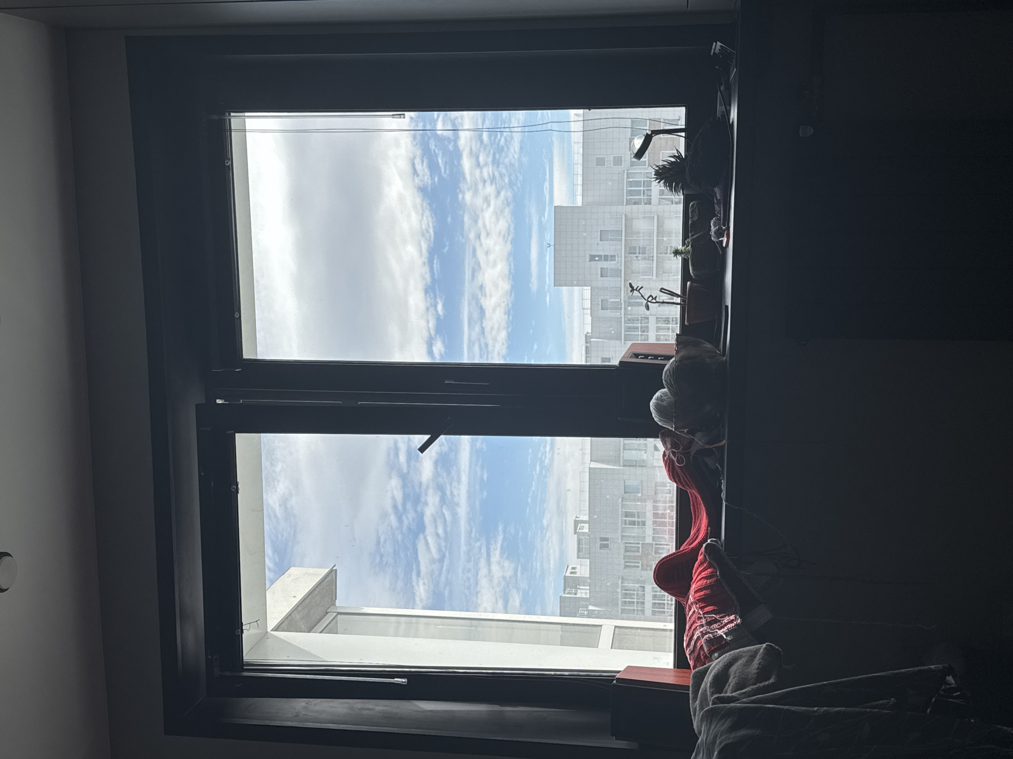 The image shows a window with a black frame. The window is divided into two sections. The upper section reveals a view of the sky with white clouds and a tall building in the distance. The lower section shows a similar view, including a rooftop structure of another building. On the window sill, there are several plants and other objects. A red knitwear and a blanket are present on the window sill. The interior is dimly lit. Based on the building style, this does not seem to be an image from Krasnoyarsk.