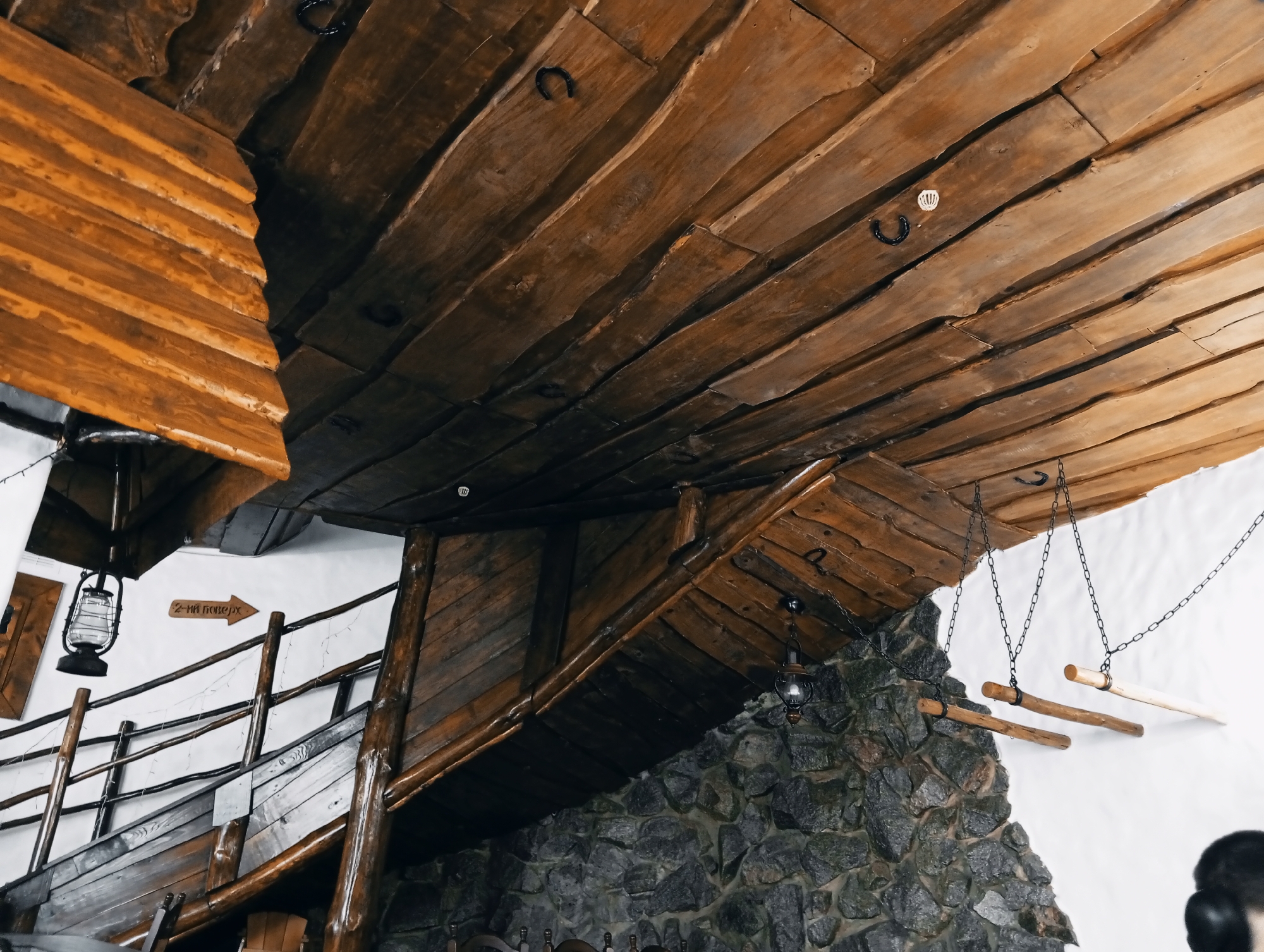 The image depicts an interior space with rustic architectural details. The ceiling is constructed from rough-hewn wooden planks, some adorned with horseshoes. A wooden staircase with a traditional design is visible, along with a stone wall. There are hanging lights and decorative elements, adding to the rustic ambiance. A sign with text in Cyrillic is visible. Based on the architectural style and available information, it is highly probable that the location is within a traditionally themed restaurant or recreational facility in Poltava, Ukraine.