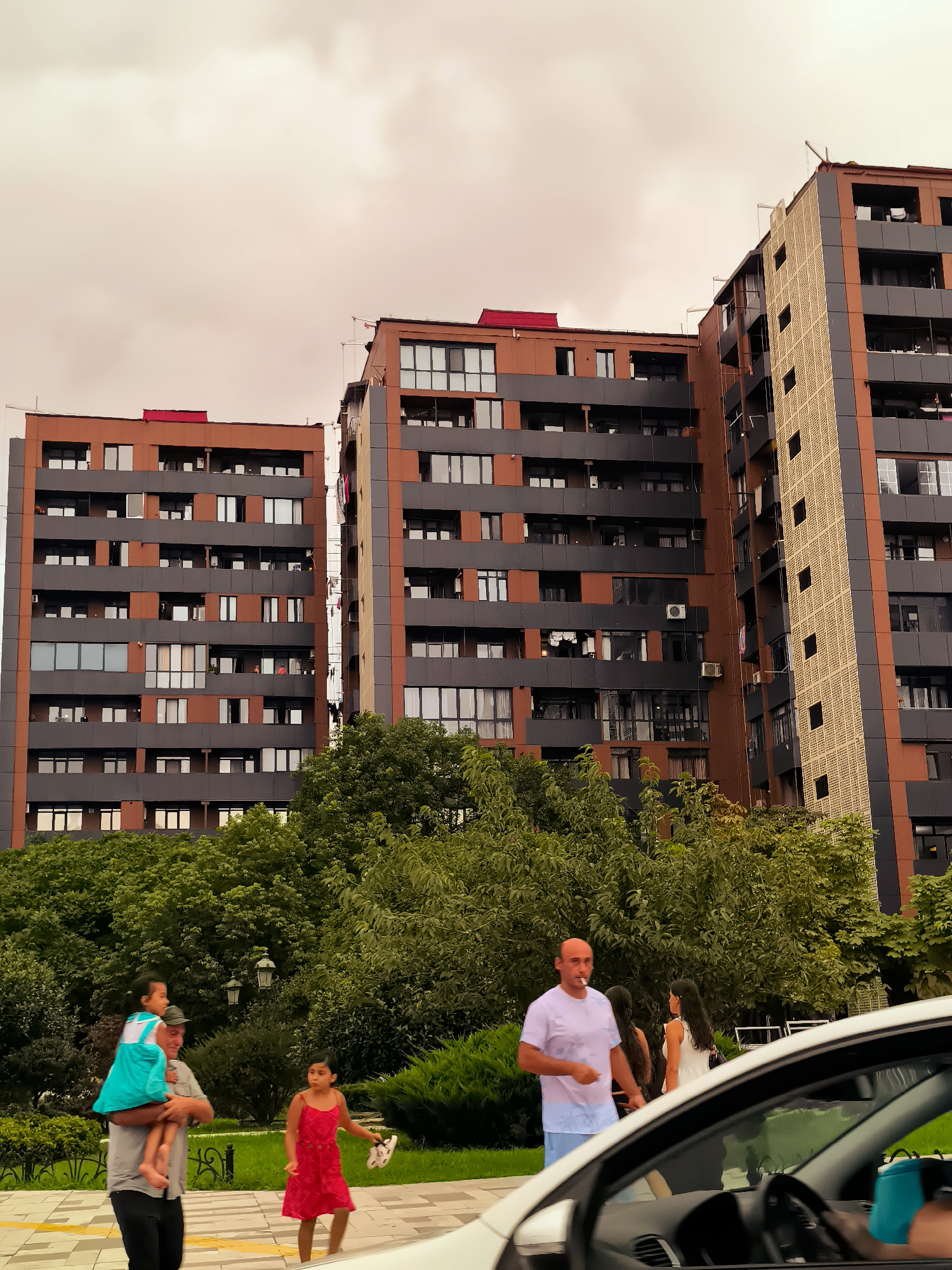 This is a photograph taken in Batumi, Georgia, featuring a modern apartment complex with distinct brown and gray facades, interspersed with patterned sections. The sky is overcast and appears reddish, suggesting either sunset or a cloudy day. In the foreground, a white car's interior is visible, with a steering wheel and dashboard elements. Outside the car, several people are walking. A man with a shaved head is smoking a cigarette, wearing a white t-shirt and light blue shorts. Behind him, two women are walking, one in a white tank top and the other with long dark hair. To the left, an older man carries a young girl on his shoulders, who is wearing a blue dress. Another young girl in a red dress walks beside them. Lush green trees and manicured bushes form a backdrop to the people, with street lamps visible amongst the greenery. The architecture of the apartment buildings suggests a contemporary urban setting.