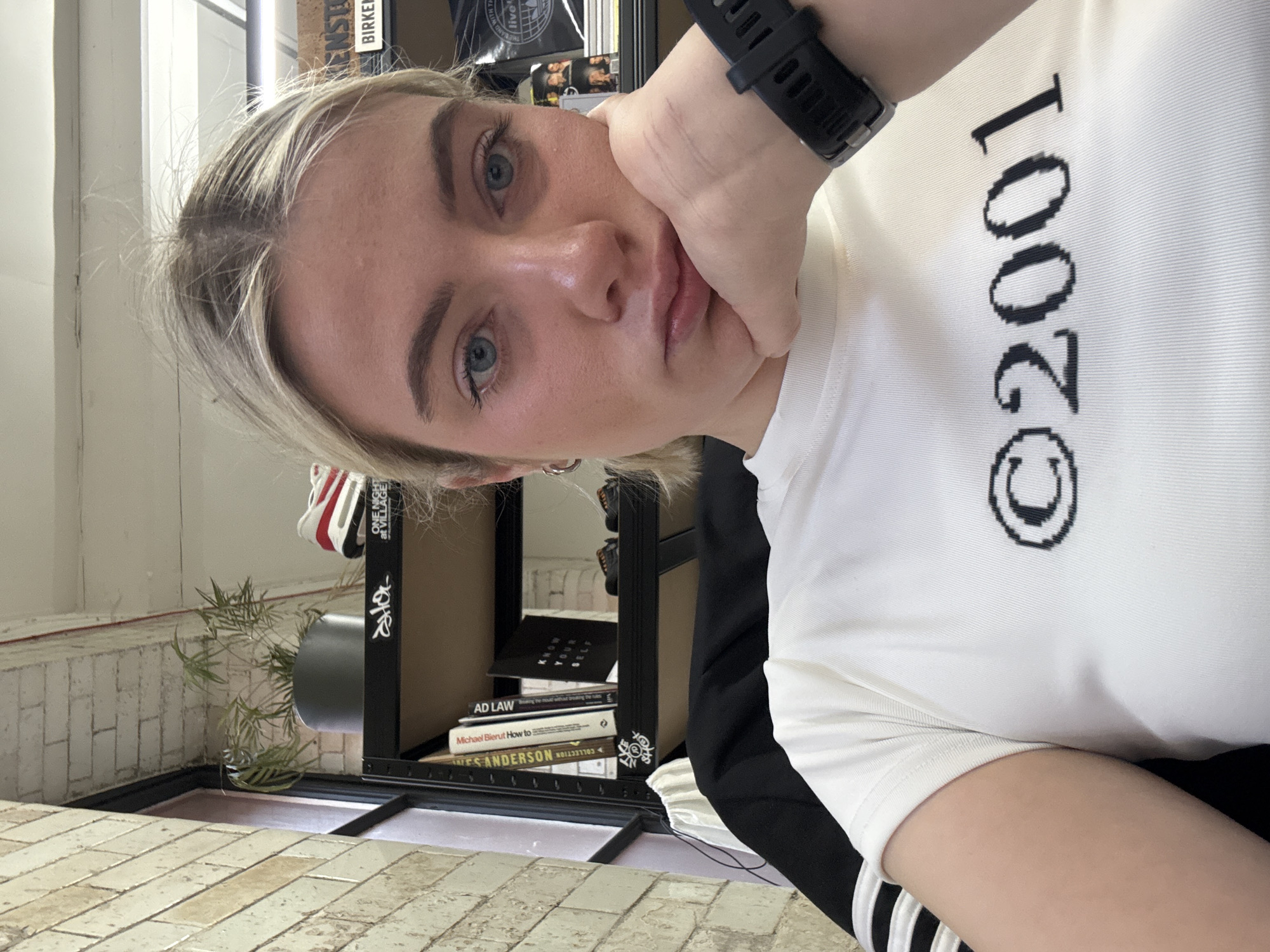 The image is an eye-level selfie, rotated slightly to the right. The subject is a young woman with light blond hair and blue eyes, resting her face on her left hand, which sports a black smartwatch. She is wearing a white t-shirt with "©2001" printed vertically in black.
In the background, a black metal shelving unit is visible, containing books, a red and white object (possibly a shoe or garment), and decorative items. Some of the book titles are discernible: "AD LAW", "Michael Bierut How to...", and "...S ANDERSON COLLECTION". A plant in a gray cylindrical pot sits on the floor near the shelving unit.
The floor is tiled with light-colored rectangular tiles. A window is visible in the upper left corner of the image. The environment appears to be indoors, possibly in a residential setting or a shop in Manchester.