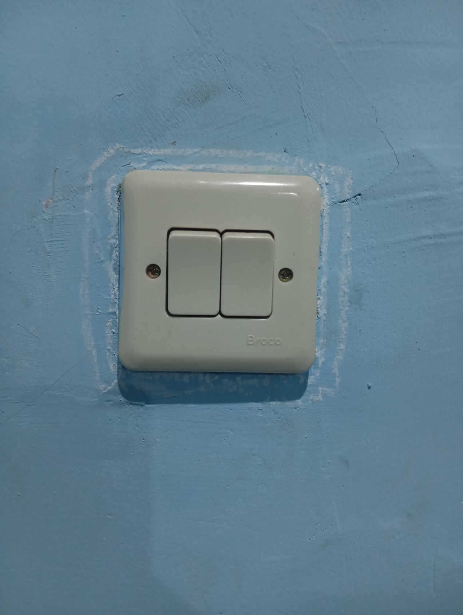 The image shows a white, double-gang light switch mounted on a light blue, textured wall. The switch plate has the brand name "Broco" printed on it. There are two rocker-style switches. The wall surface surrounding the switch plate appears uneven and has some white chalk-like markings, suggesting it might have been used for marking measurements or outlines. There are also visible cracks and imperfections in the wall. The lighting in the image creates subtle shadows around the switch plate. This light switch is a common electrical fixture found in homes and buildings. In the context of Sudikampiran, Indonesia, such a switch would likely be found in a residential dwelling or a public building, contributing to the basic infrastructure of the area.