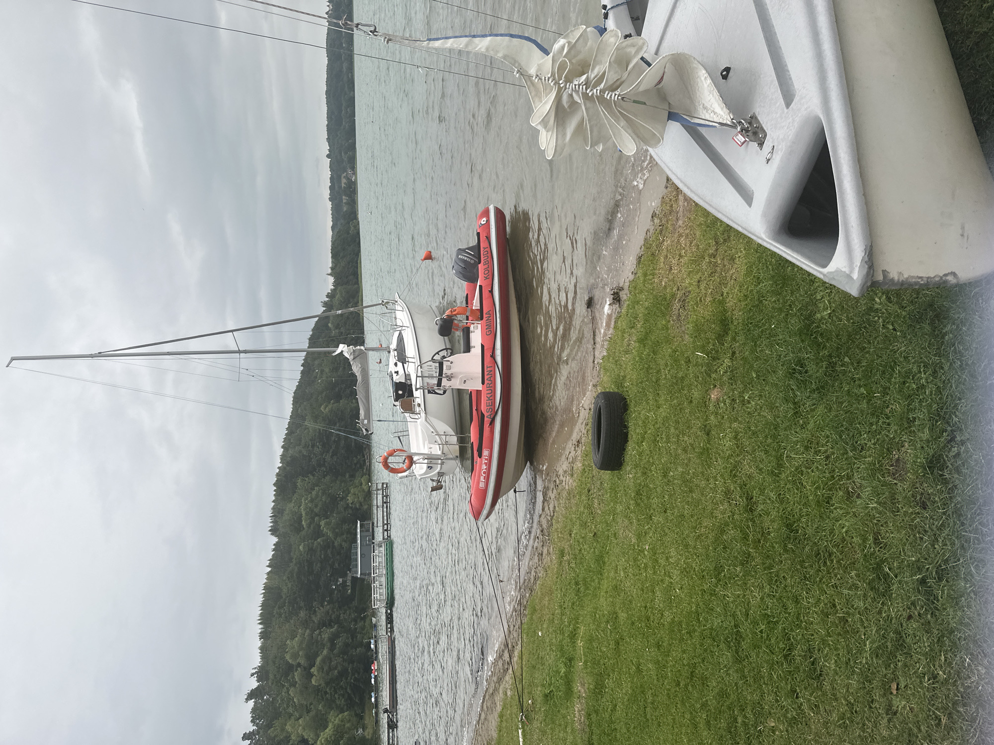 An overcast day at a lake in Chmielno, Poland, features a shoreline with grass and a sandy edge. Moored boats are visible on the water, including a white sailboat with a mast, a red and white inflatable rescue boat with "ASEKUKANT" and "KOLBUDY" written on it, and a portion of a small gray sailboat in the foreground. The rescue boat has an outboard motor and a life preserver attached to its side. A deflated black tire rests on the grass near the water's edge. In the background, a tree-lined shore with some structures on a dock is visible.