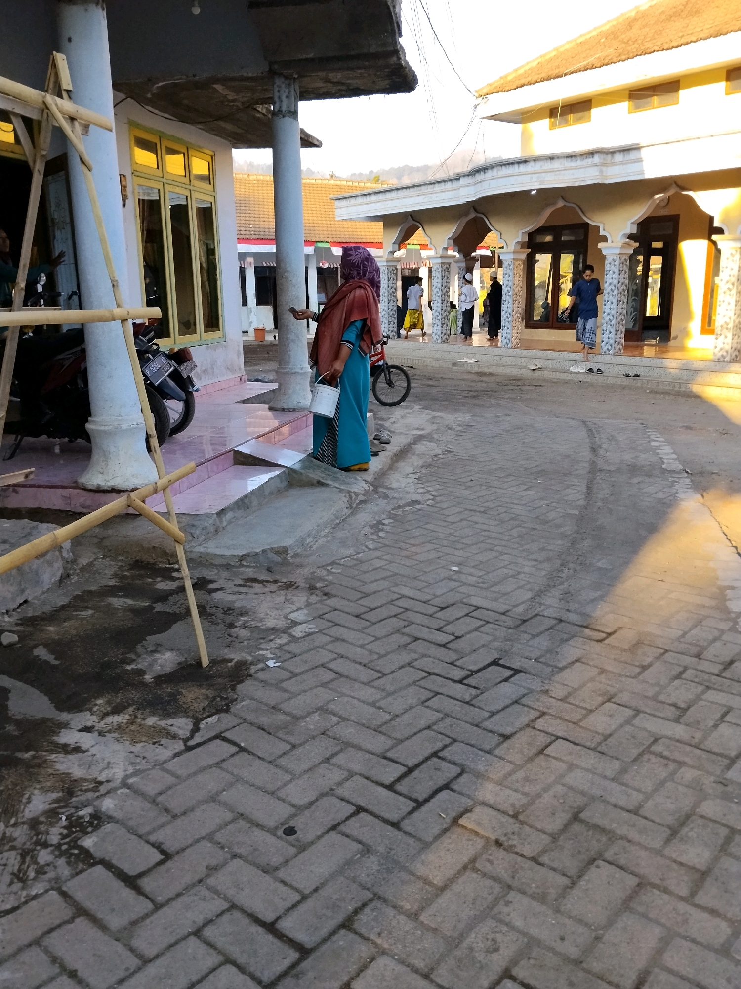 This image depicts an outdoor scene in Panggungan, Indonesia, likely in front of a mosque or a similar community building. The foreground is dominated by a paved walkway made of interlocking gray bricks, with some wet patches and debris visible. To the left, a motorcycle is parked near a white pillar supporting a roofed structure with yellow-trimmed windows. Bamboo scaffolding is also present on the left side of the image, suggesting ongoing construction or maintenance.
In the mid-ground, a person wearing a maroon headscarf and a blue garment is standing, holding a white bucket and a mobile phone. Adjacent to this person is a bicycle. Further into the scene, people are seen walking and standing near the entrance of a building with arched colonnades and yellow trim. Several men are visible, some wearing traditional Muslim attire. The building itself has a tiled roof and appears to be a significant communal structure. The lighting suggests it is either early morning or late afternoon, with warm sunlight casting shadows. The environment appears to be a built-up area with clear skies and a distant view of hills.