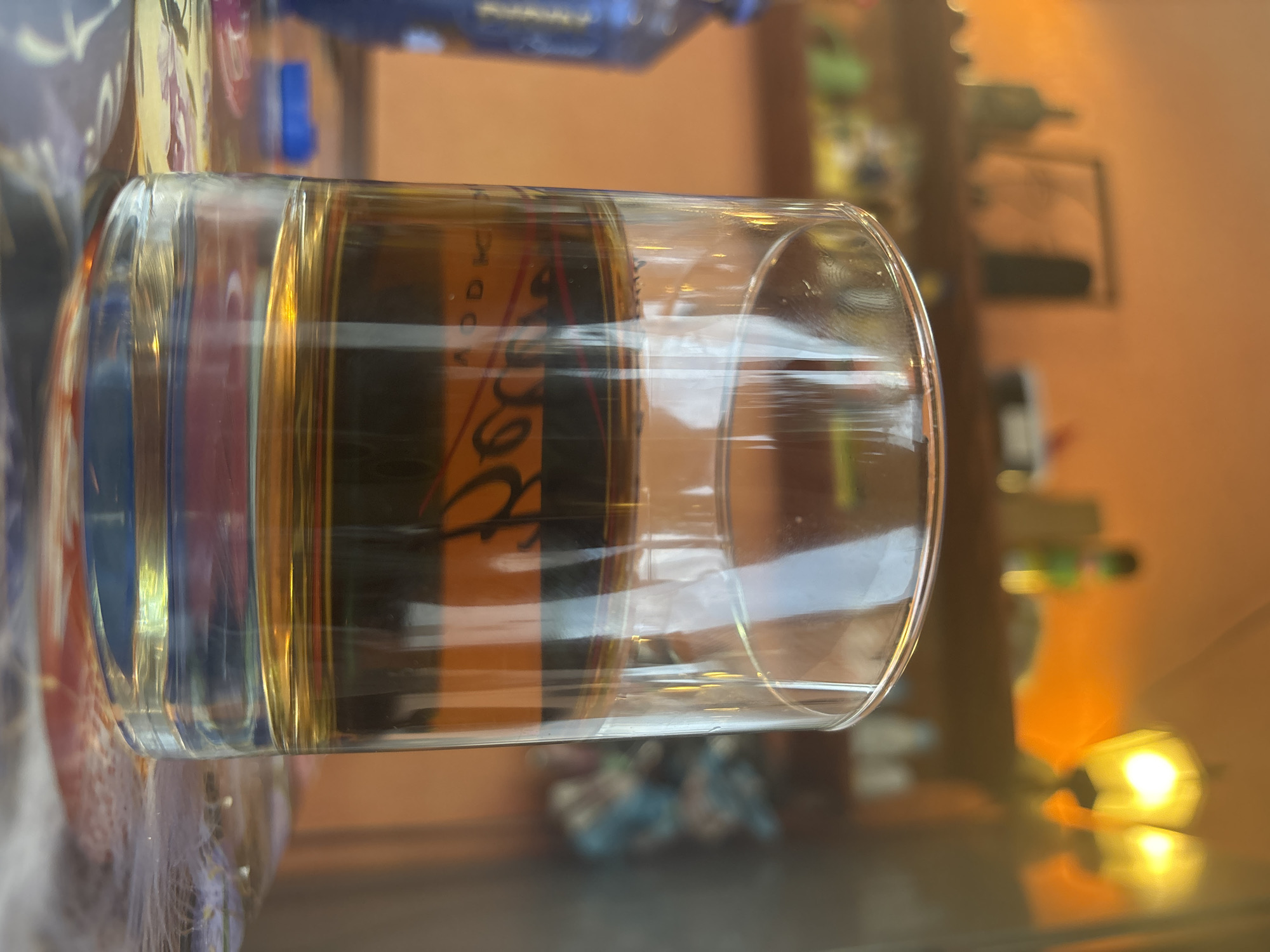 A close-up, slightly out-of-focus photograph shows a glass of amber liquid placed on a reflective surface. The side of the glass has a label with the word "Rogo" visible. A partially visible bottle is in the upper background. Beyond the reflective surface, a cluttered interior includes indistinct objects and light sources, suggesting a domestic or commercial setting. No people are visible. The location, Lom, Bulgaria, is not discernible from the image.