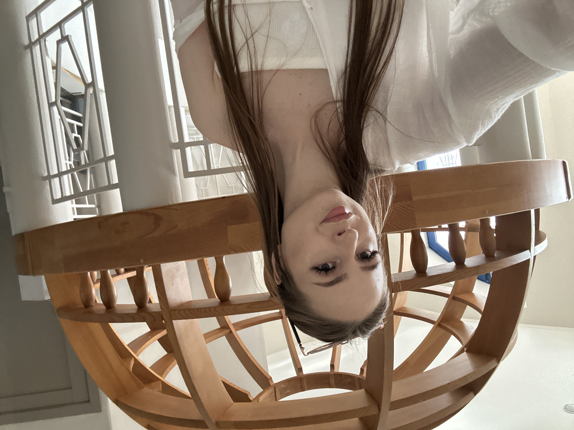 A young woman with long brown hair and wearing a white tube top and a sheer white shirt is taking a selfie in front of a large wooden dome structure. She has on false eyelashes and light makeup, and sunglasses are perched on her head. The background appears to be an indoor space with white walls and windows with white decorative grilles.
