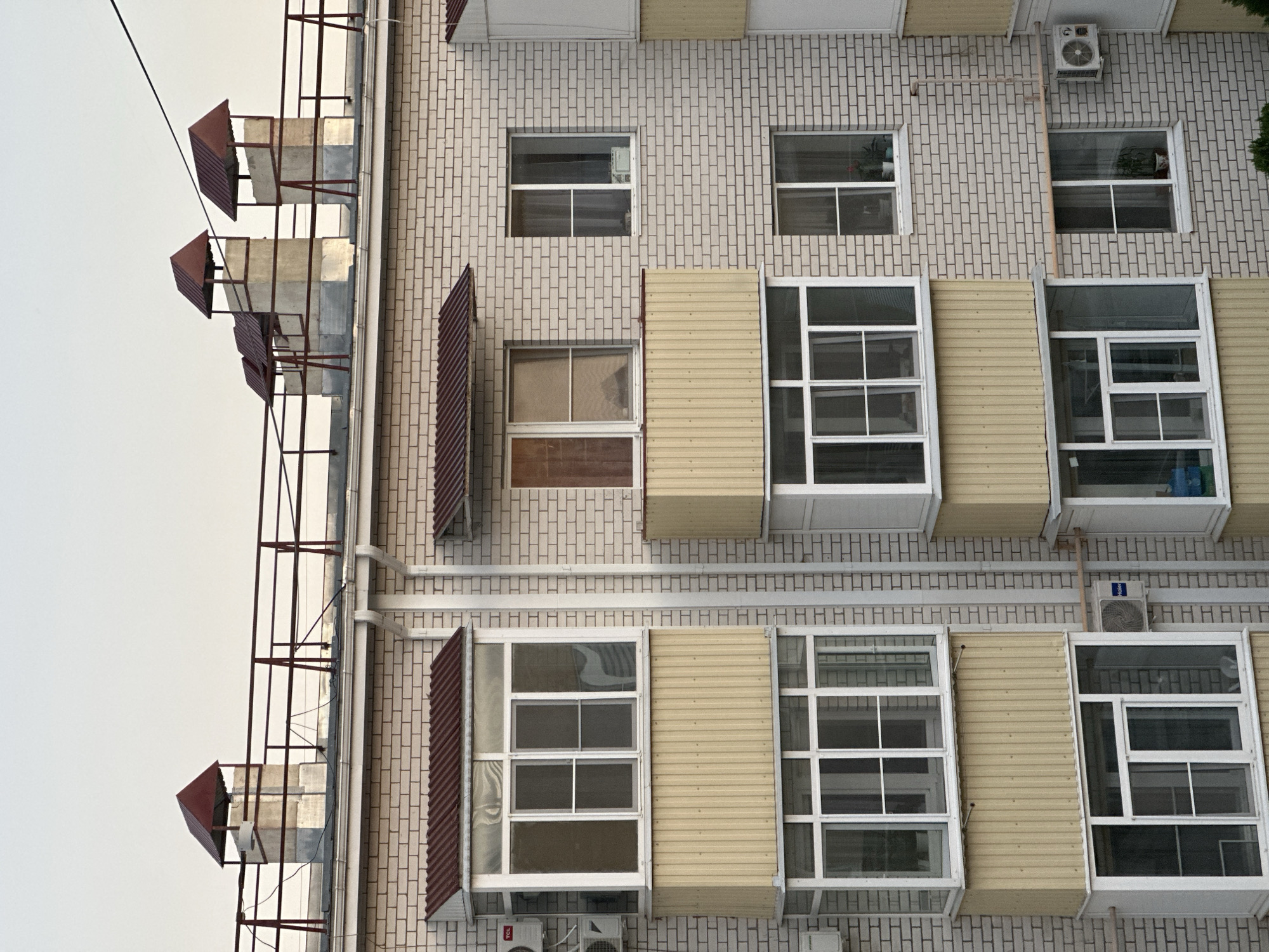 The image is a close-up, high-angle exterior view of a multi-story apartment building constructed with light-colored brick. The building features numerous windows and balconies. Some balconies are enclosed with glass, while others have yellow corrugated metal siding. The roofline of the building is visible, topped with several rectangular brick chimneys, each capped with a red metal roof. A red metal railing runs along the edge of the roof. On the building's facade, there are several air conditioning units mounted on the walls. The sky is clear and appears to be late afternoon or early evening. The scene is devoid of people or any observable activity. The image has been identified as being taken in the city of Nevinnomyssk, Russia.