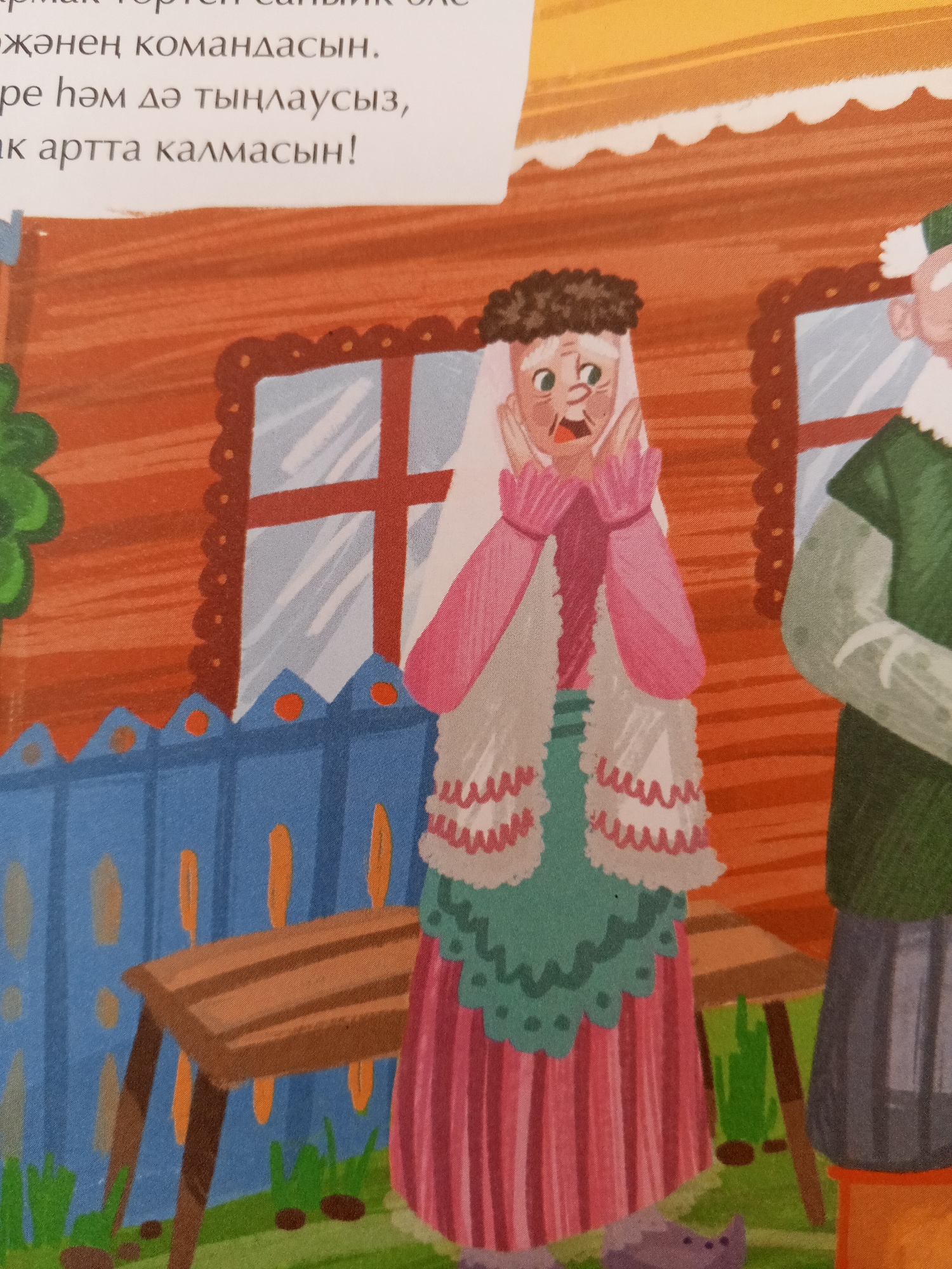 This image depicts a scene with two characters in an outdoor setting, possibly a village or rural area, given the rustic fence and wooden table. The text above the image appears to be in a non-Latin script, likely Cyrillic or a related alphabet, suggesting a Russian context. The left character is an older woman with white hair, wearing a pink dress with a patterned trim and a white head covering. Her hands are raised to her face in an expression of surprise or concern. To the right is an older man wearing a green tunic and a white cap. He stands with his arms folded. The background shows a wooden structure, possibly a house, with a window. The overall style is illustrative, suggesting it might be from a children's book or a similar publication. There is no specific information within the image to pinpoint a location within the city of Chistopol, Russia.